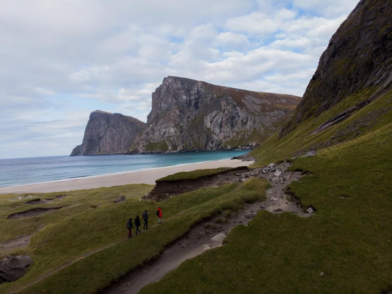 Four hikers walk along a grassy path toward a sandy beach, bordered by steep green hills and dramatic rocky cliffs, under a mostly cloudy sky—capturing moments for photos to be posted later.