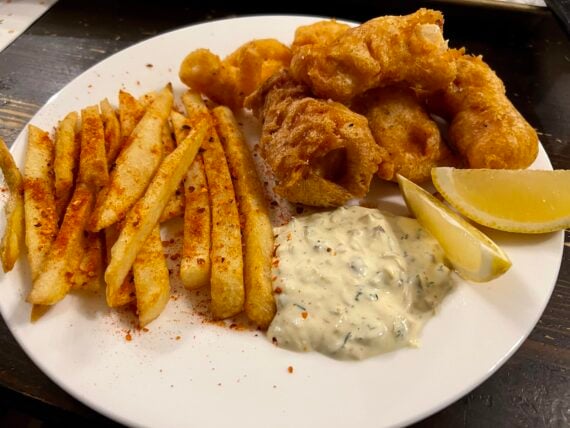 A plate of crispy fries with seasoning, battered fried fish, tartar sauce, and two lemon wedges on a white plate.