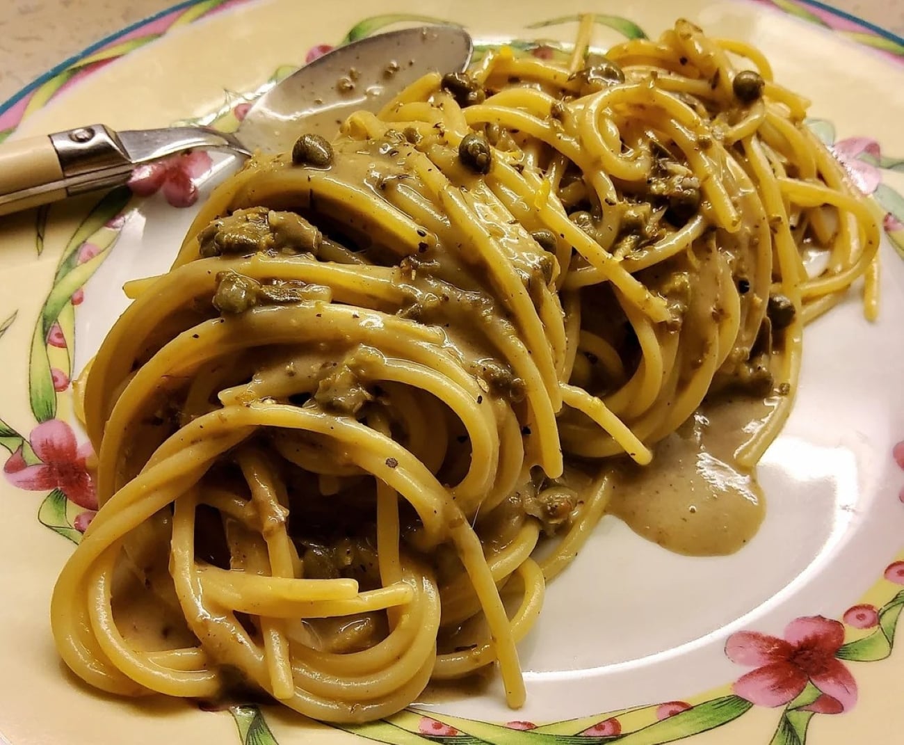 Spaghetti topped with a creamy, pepper-speckled sauce and capers is artfully presented on a floral-patterned plate with a spoon beside it, capturing the essence of classic Italian comfort in these stunning photos of pasta.