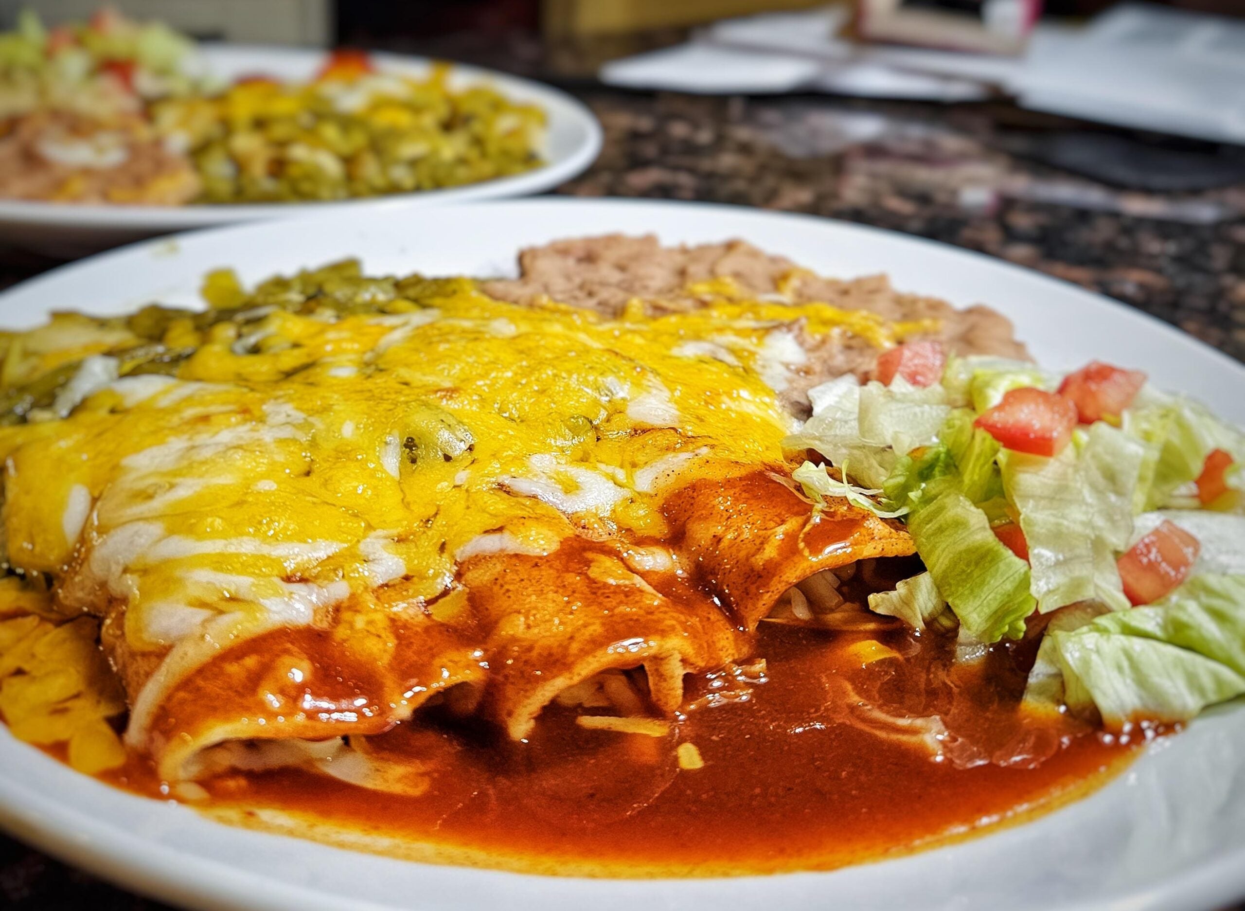 A plate of cheesy enchiladas topped with red sauce, served with refried beans and a side of lettuce with diced tomatoes. Another plate of food is blurred in the background.