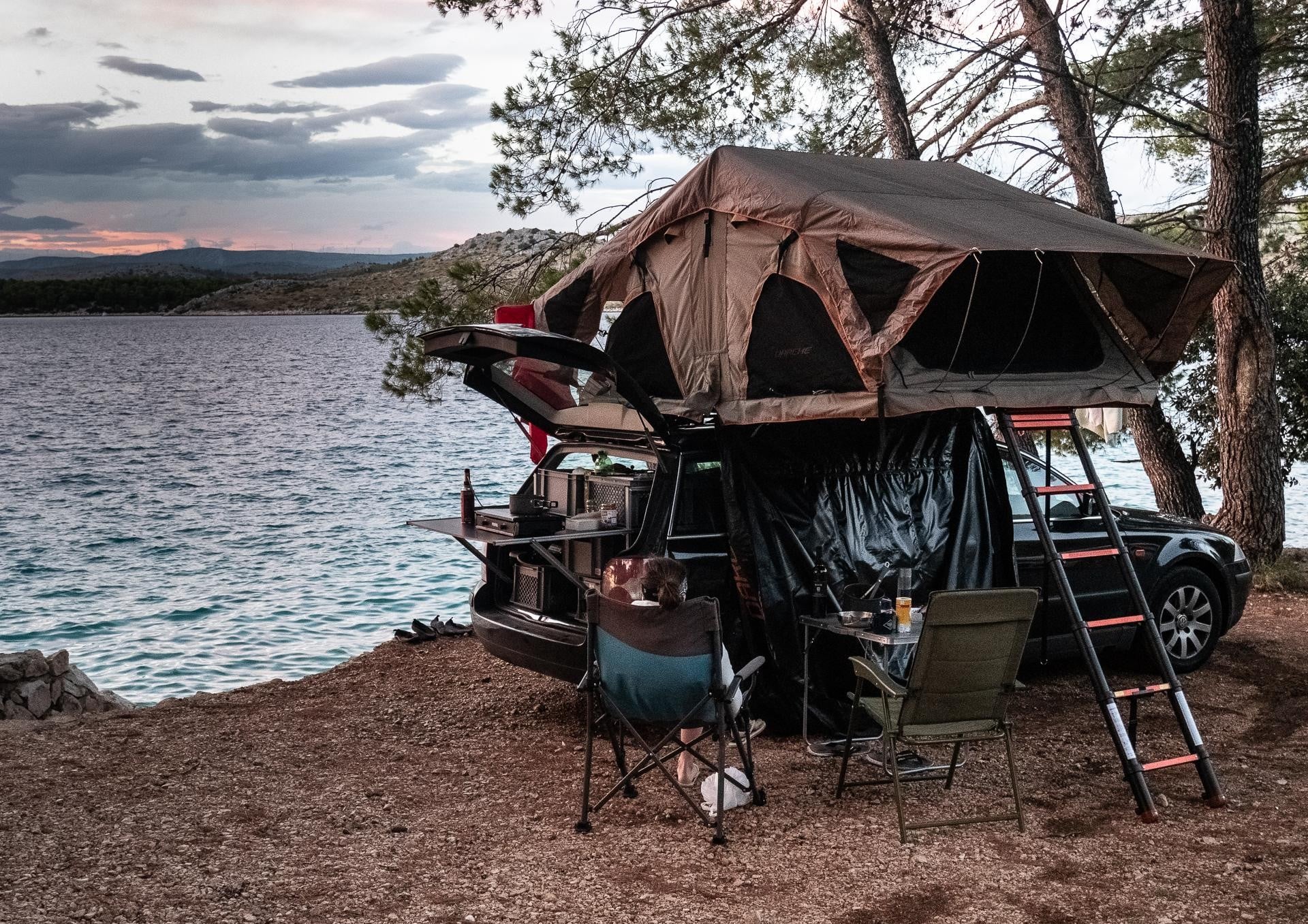 A car with a rooftop tent is parked by a lake, surrounded by trees. Camping gear, chairs, and a table are set up next to the car at sunset.