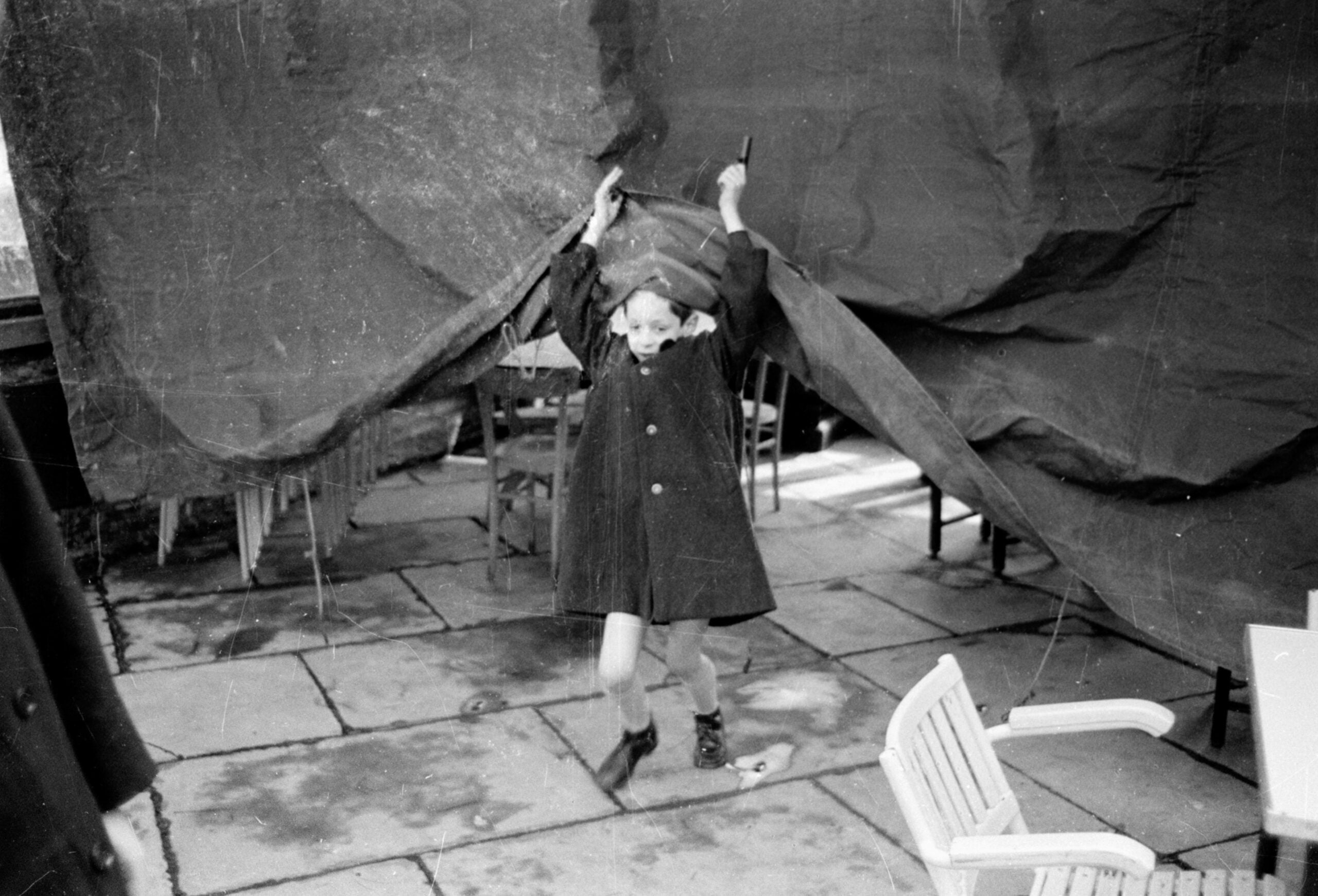 A child in a coat and boots lifts a large tarp or sheet outdoors, with scattered chairs and icicles visible in the background. The scene appears casual and candid.