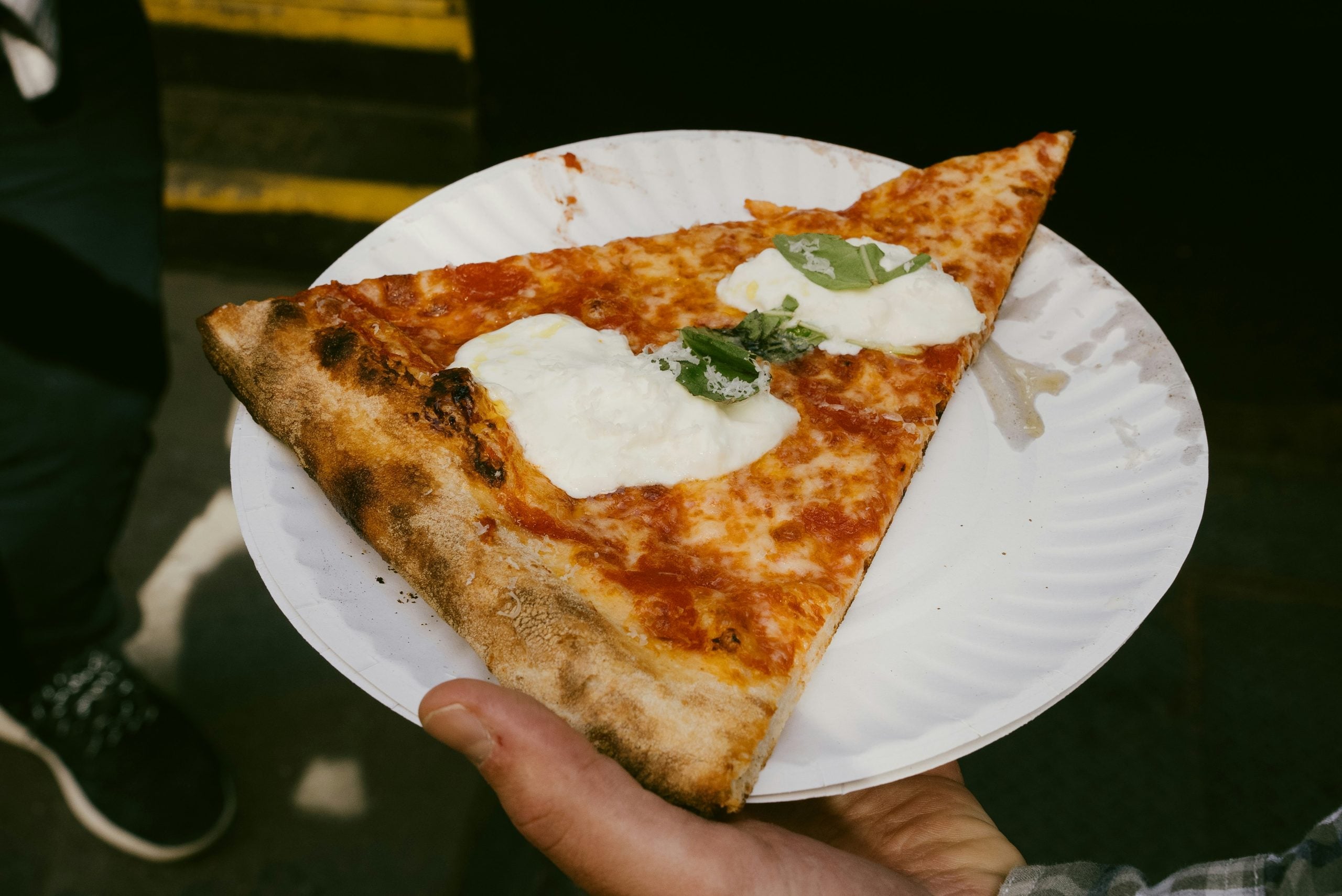 A hand holds a large slice of pizza on a white paper plate. The pizza has melted mozzarella, basil leaves, and a slightly charred crust. The background is out of focus, showing part of someone’s shoe and pavement.
