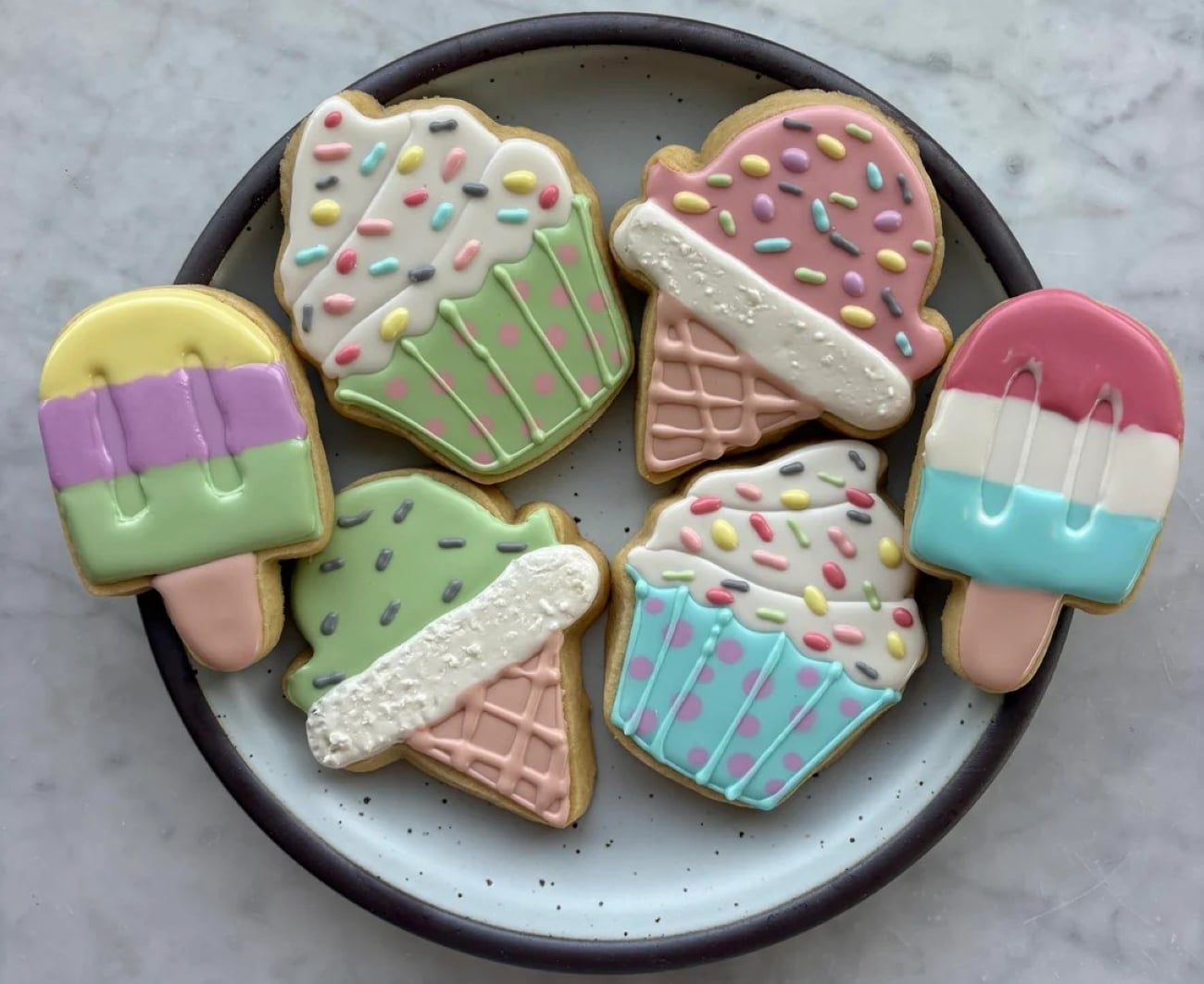 A plate of six cookies decorated as ice cream cones and popsicles, with pastel-colored icing and sprinkles, placed on a marble surface.