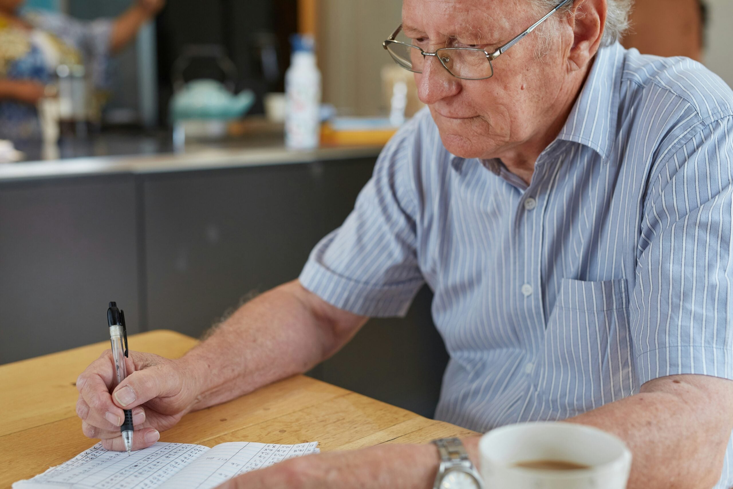 An elderly man wearing glasses and a striped shirt sits at a table, writing on a piece of paper with a pen—perhaps reviewing his retirement savings. A cup of coffee is nearby. The background shows a kitchen area out of focus.