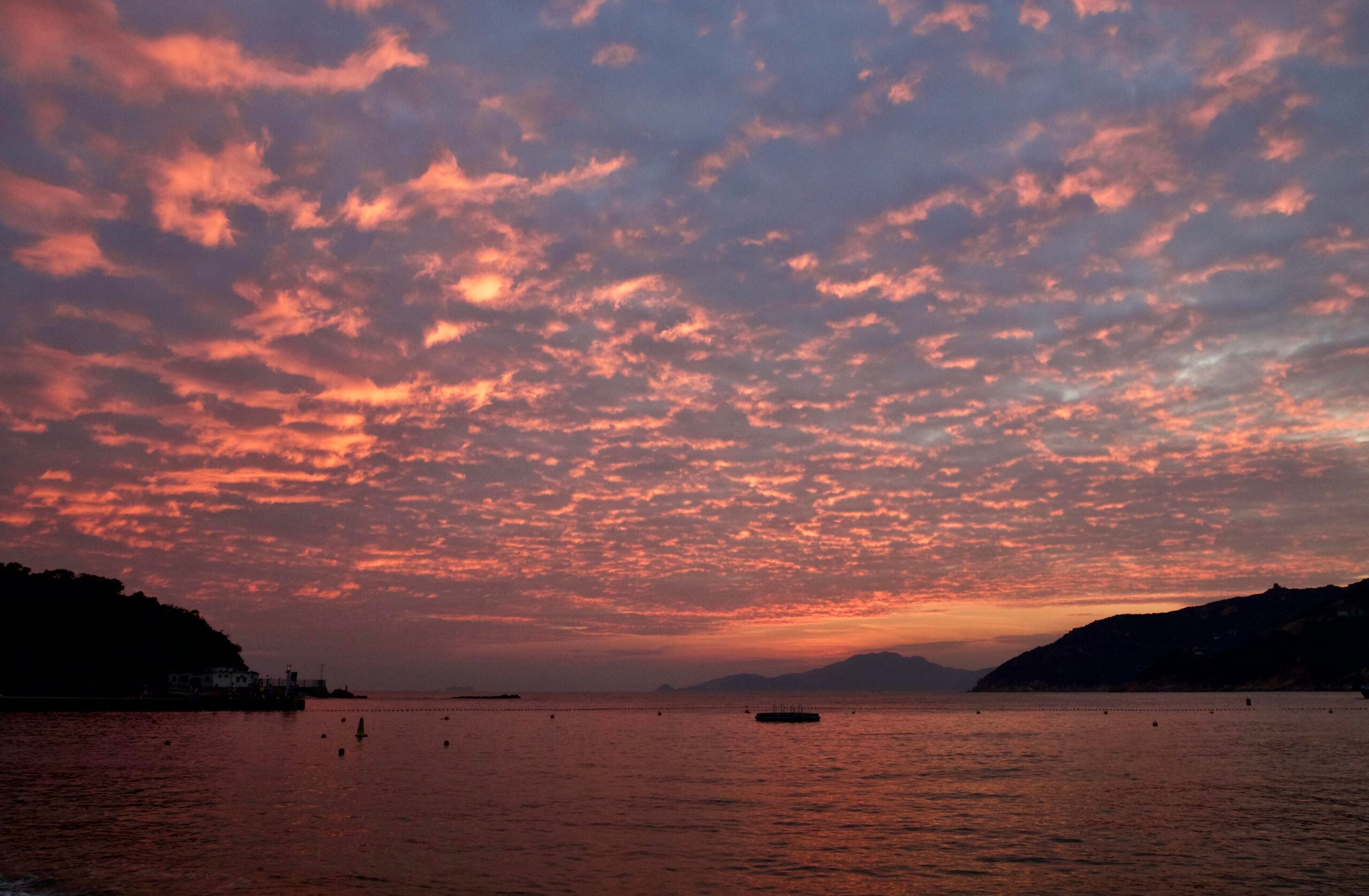 Sunset over calm water with mountains on each side, a building on the left, and a dramatic sky filled with pink and orange clouds.