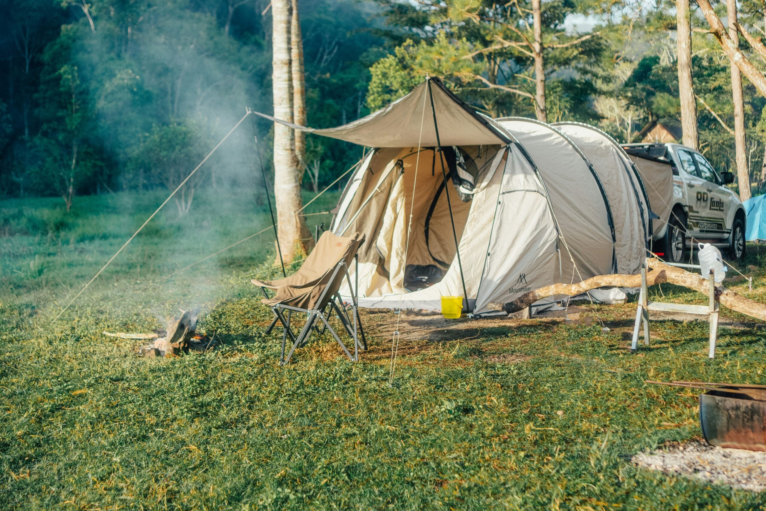 A beige tent and camping chair sit on grass near a smoking campfire, with a white car parked beside them. Trees and sunlight surround the camping area in a peaceful outdoor setting.