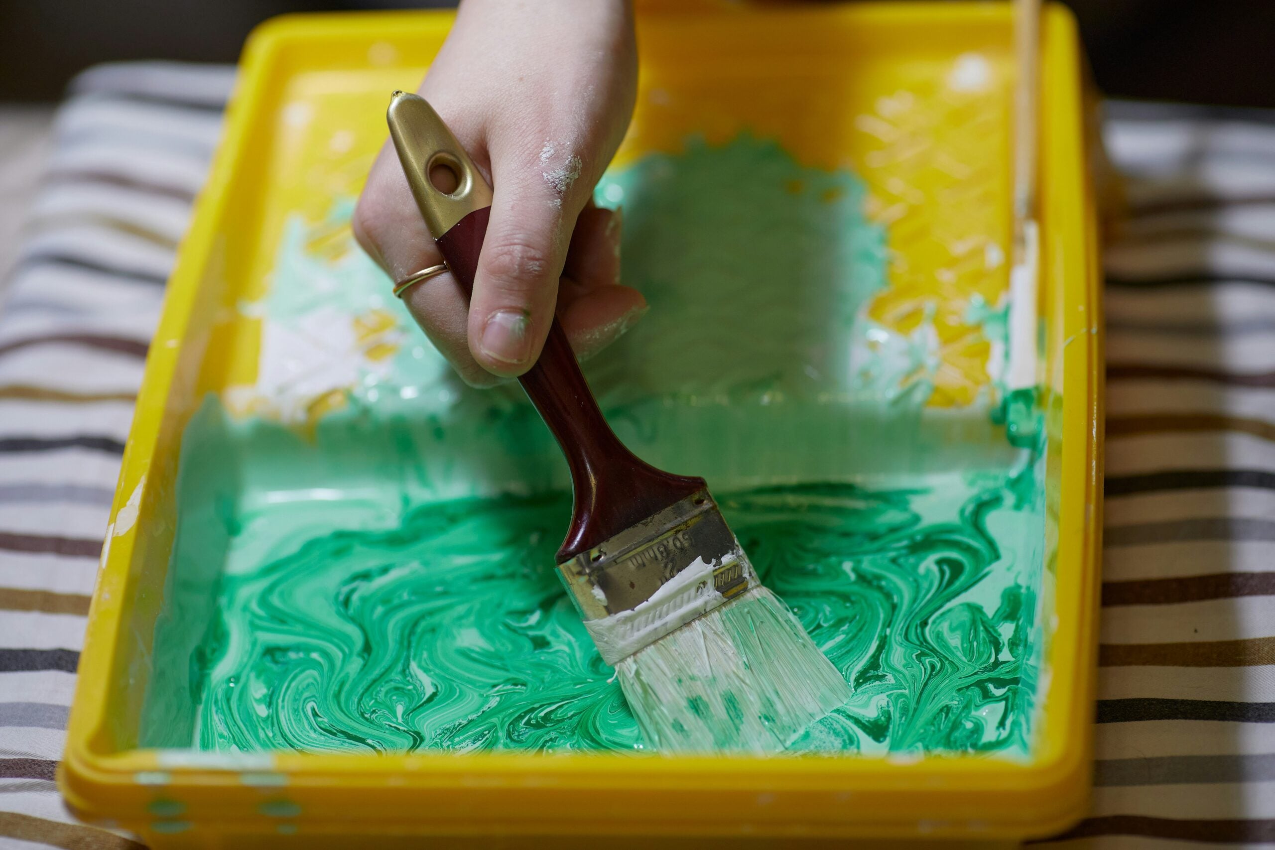 A hand holding a paintbrush is dipping it into swirled green and white paint in a yellow tray, with a striped cloth underneath.