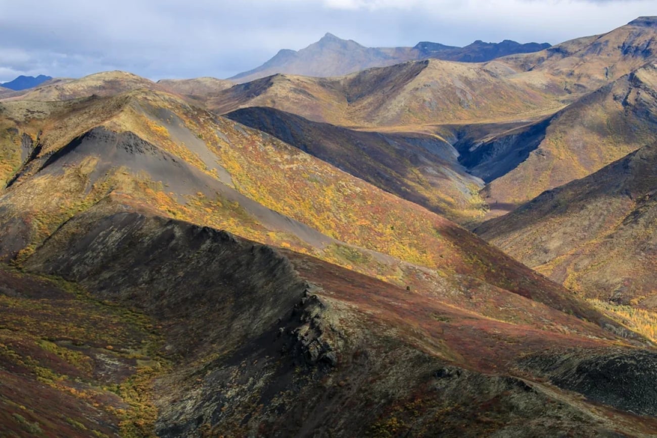 Rolling, rocky mountains with patches of yellow and red vegetation under a cloudy sky. The rugged terrain features ridges, valleys, and distant peaks, creating a sense of vast, remote wilderness.