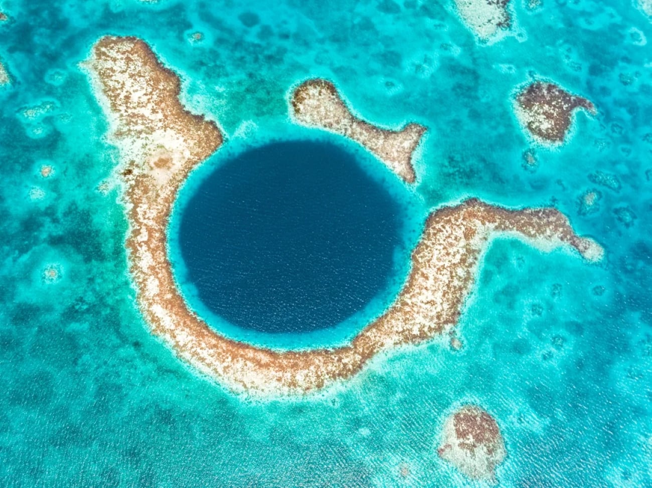 Aerial view of the Great Blue Hole in Belize, one of the most stunning tropical locations, featuring a large, circular, deep blue sinkhole surrounded by light turquoise coral reefs and shallow water.