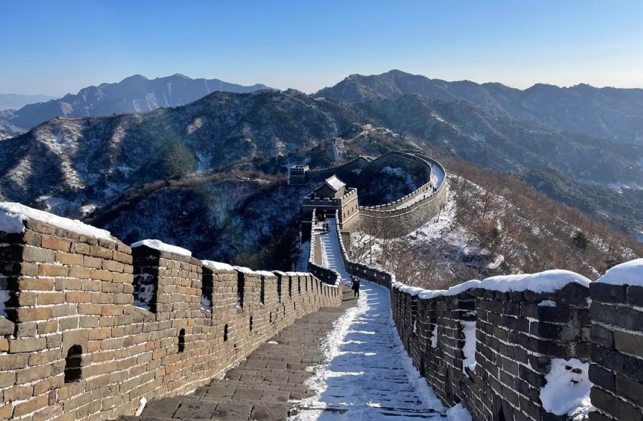A snow-dusted section of the Great Wall of China winds through mountains under a clear blue sky, with sunlight casting shadows on the ancient stone structure.