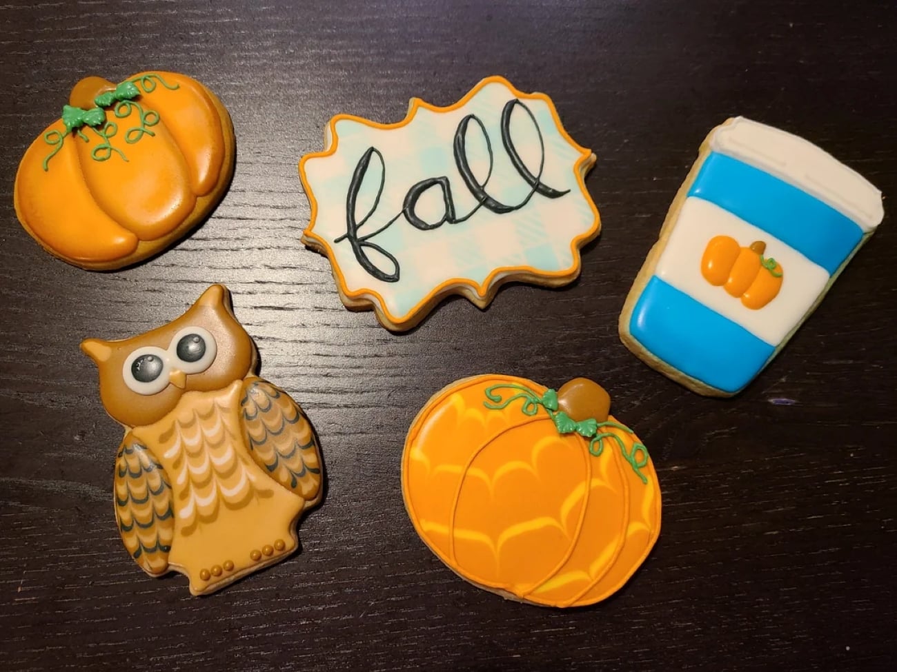 Decorated cookies shaped like pumpkins, an owl, a coffee cup with a pumpkin, and a plaque with the word “fall” on a dark wooden surface.
