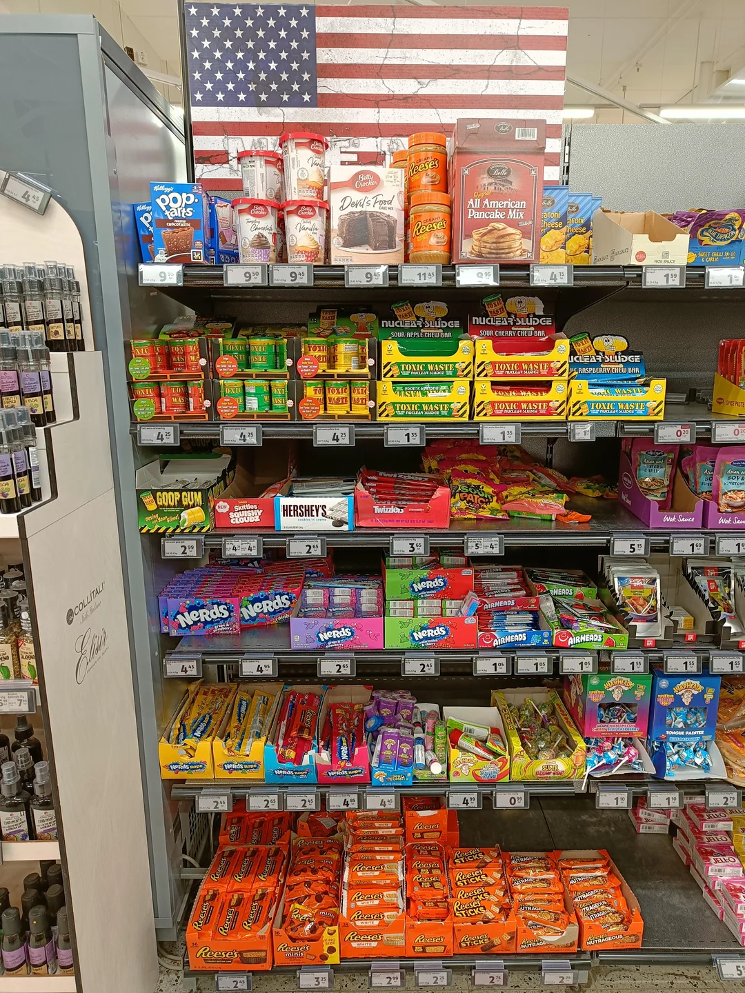 Shelves in a store filled with assorted American snacks and candies, including Reese’s, Nerds, and Pop-Tarts, with a large American flag displayed at the top.