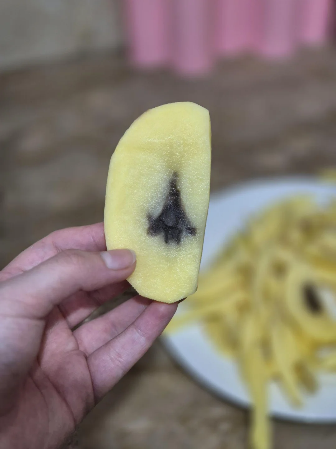 A hand holds a peeled slice of mango with a dark discoloration in the center resembling a spade or tree shape. A plate with more mango slices is blurred in the background.