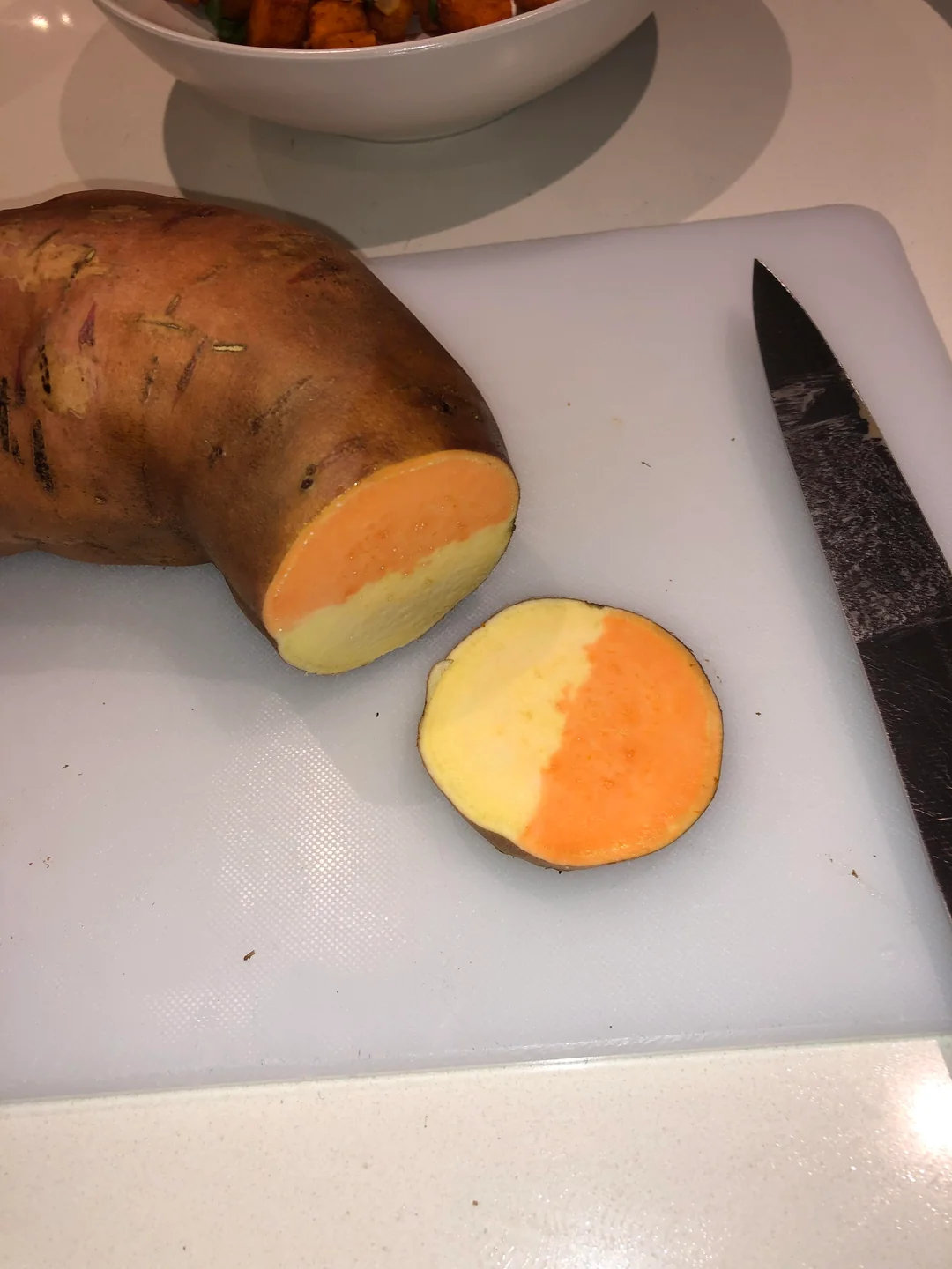 A partially peeled sweet potato with both orange and white flesh, sitting on a cutting board next to a knife. One round slice shows the distinct color split.