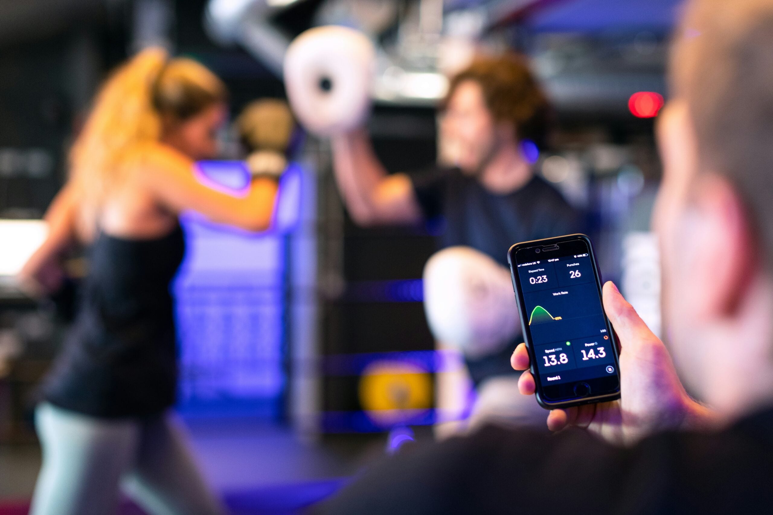 A person holds a smartphone tracking stats while two people spar in a boxing gym, one with gloves raised. The background is blurred, focusing on the phone display.