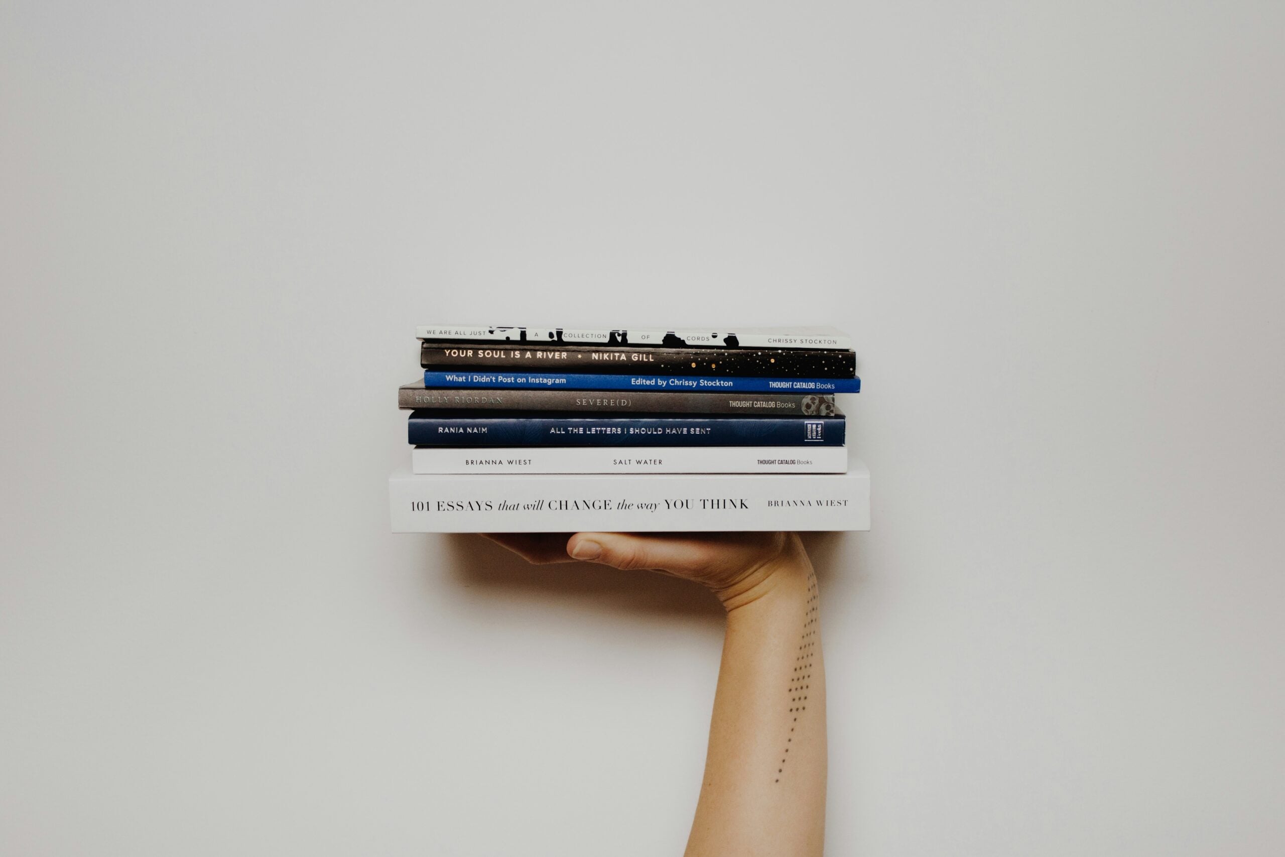 A hand holds a stack of five books against a plain white background. The forearm has a vertical line of small text tattooed on it. The books are stacked horizontally, with the largest book at the bottom.