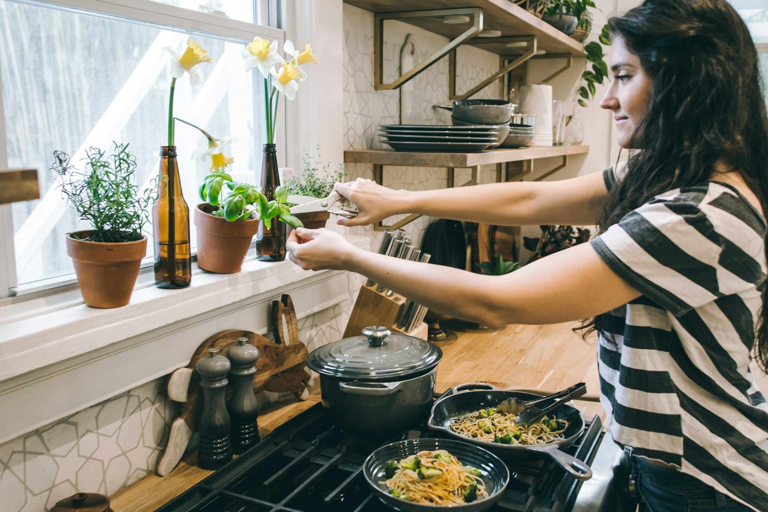 A person in a striped shirt chops fresh herbs and sprinkles them on pasta in a kitchen with plants, flowers, and dishes by the window.