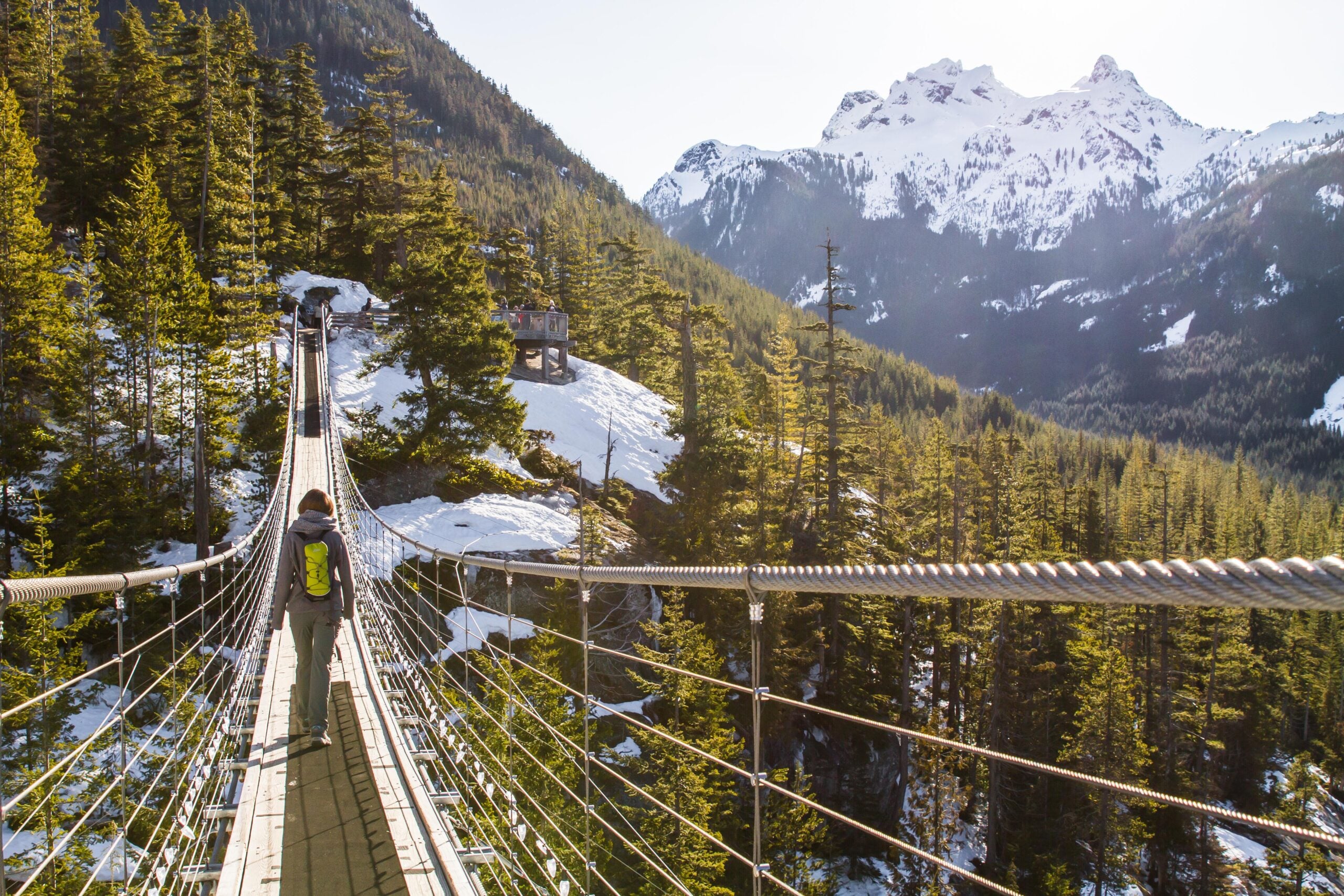 A person with a green backpack crosses a long suspension bridge surrounded by evergreen trees, with snow-capped mountains visible in the background under a bright, clear sky.