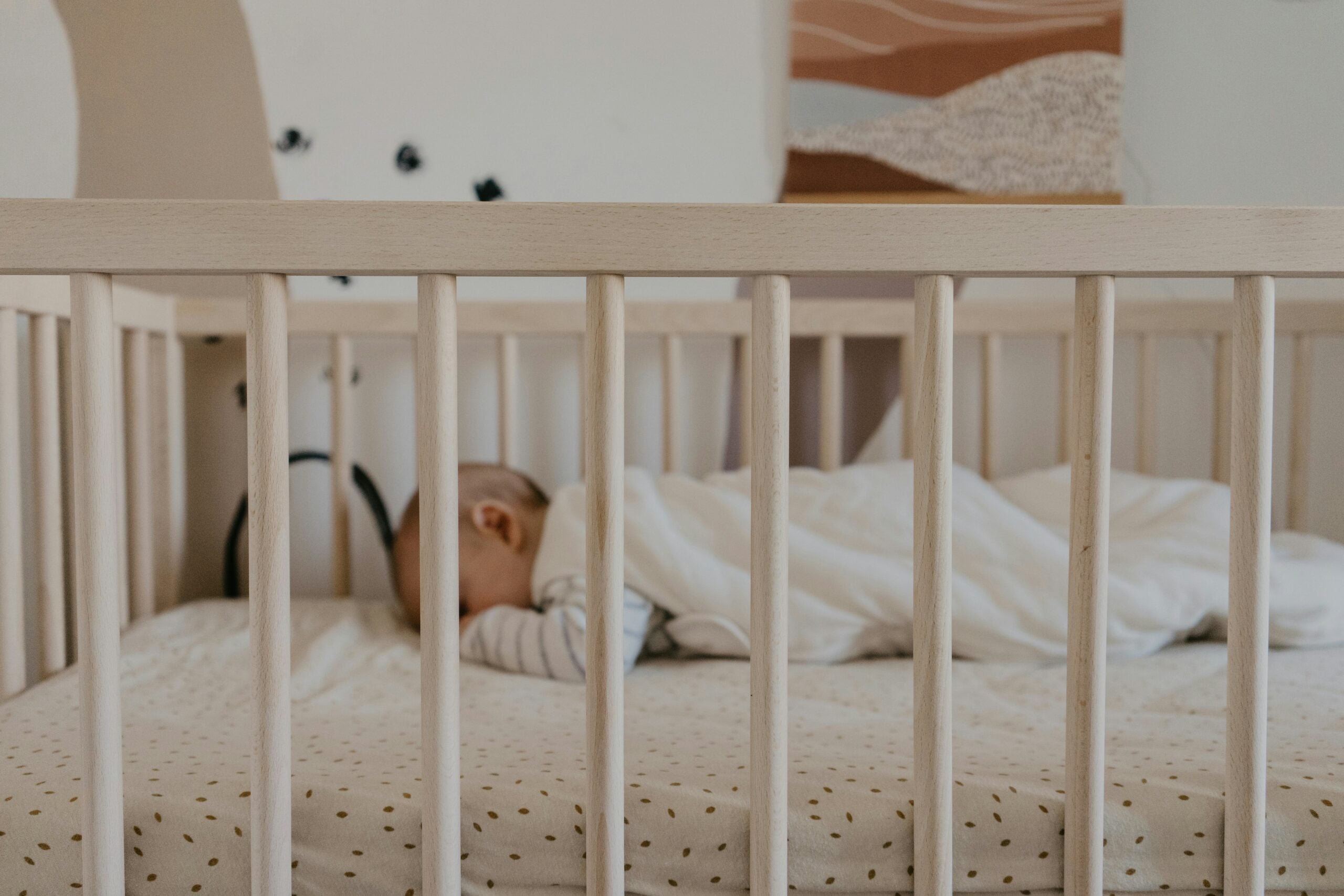 A baby sleeps on their stomach in a wooden crib with a white blanket. The crib bedding is cream with gold dots. The background features soft, neutral decor and abstract wall art.