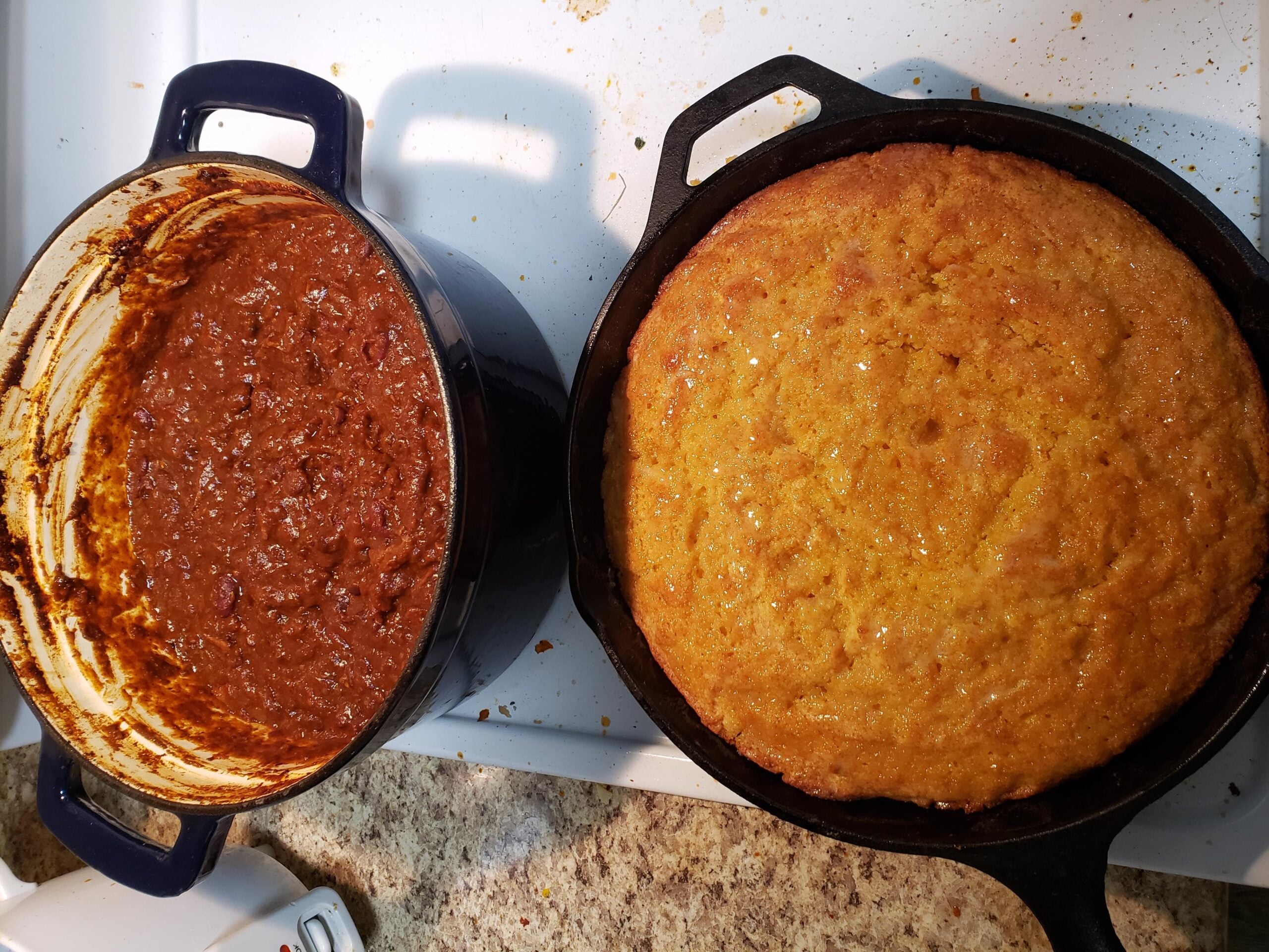 A pot of chili sits next to a skillet of freshly baked cornbread on a kitchen counter, both in cast iron cookware.