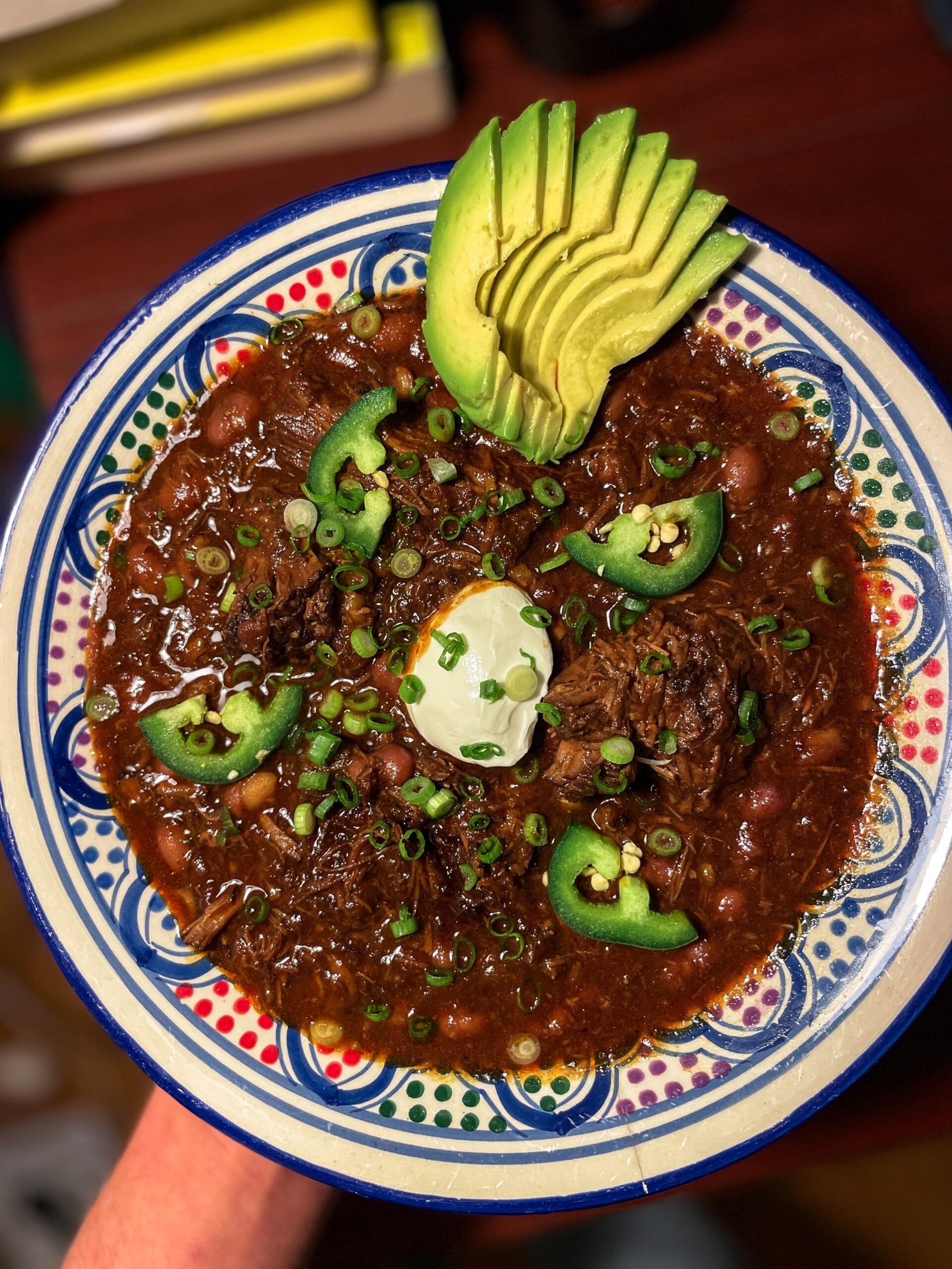 A colorful bowl of chili topped with sliced avocado, green onions, jalapeño slices, and a dollop of sour cream, served on a patterned plate.