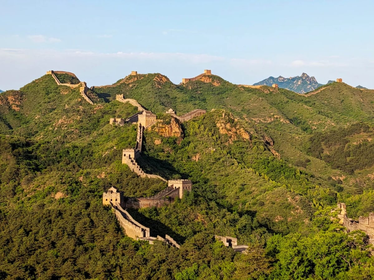 The Great Wall of China winds over green, tree-covered hills under a clear blue sky, with watchtowers visible along its length.
