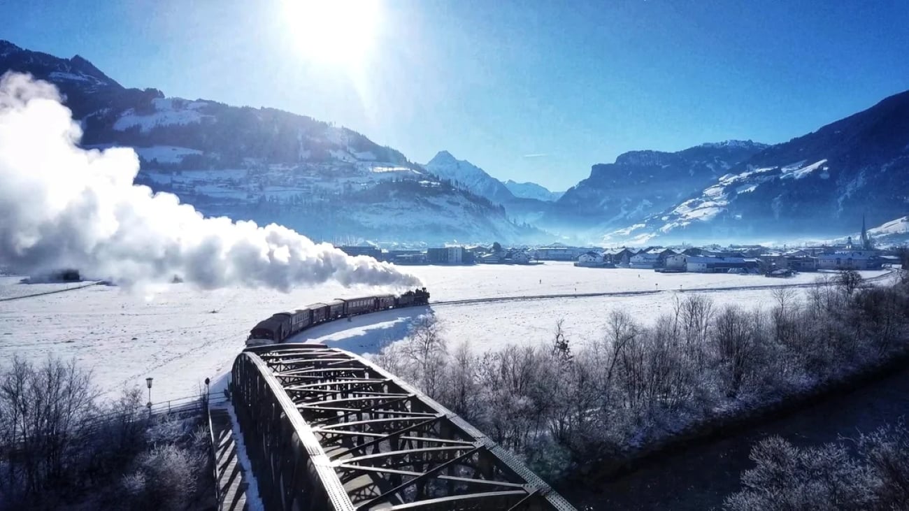 A train crosses a metal bridge over a river, trailing steam through a snowy valley surrounded by sunlit mountains under a clear blue sky.