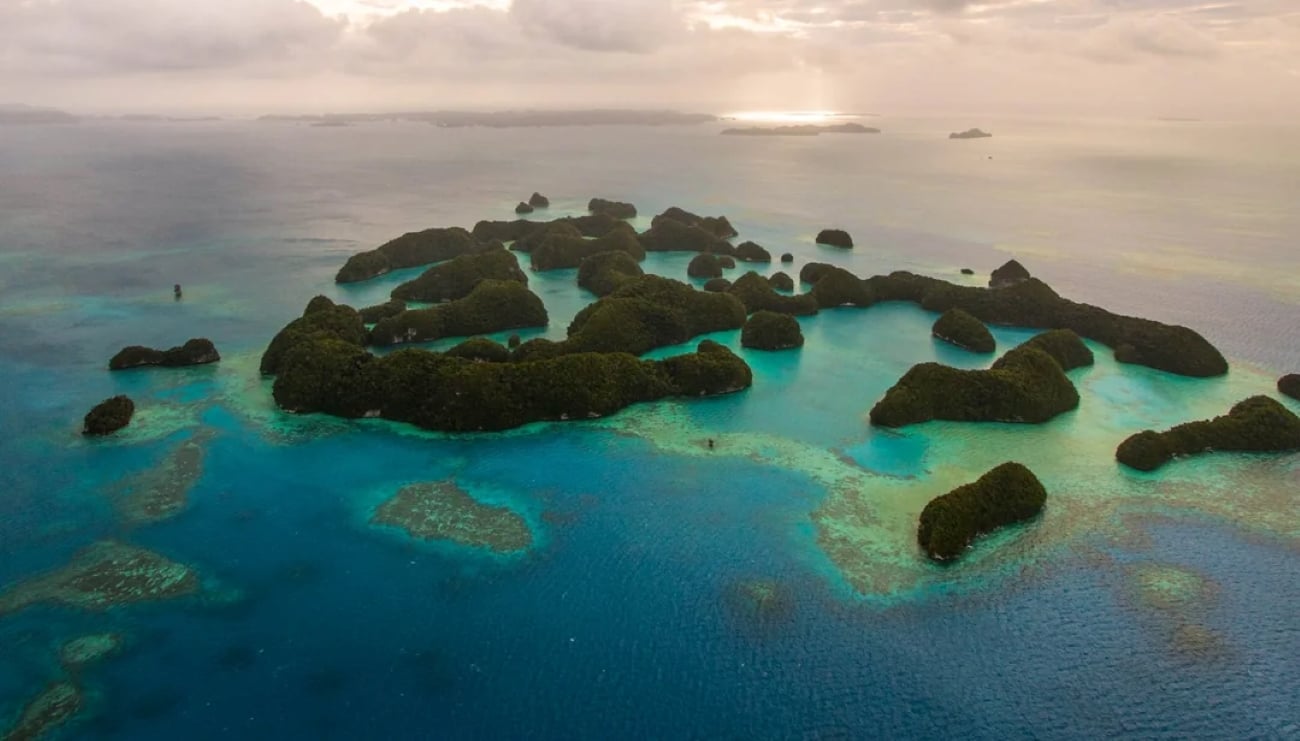 Aerial view of small, lush green islands in tropical locations, surrounded by turquoise and deep blue ocean waters under a cloudy sky with sunlight peeking through.