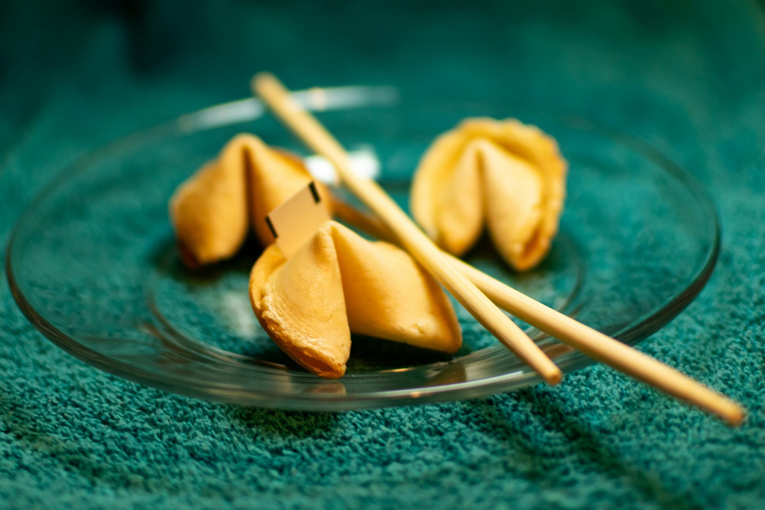 Three fortune cookies and a pair of wooden chopsticks are arranged on a glass plate, placed on a teal textured surface. One cookie is cracked open, and a fortune slip is partially visible.