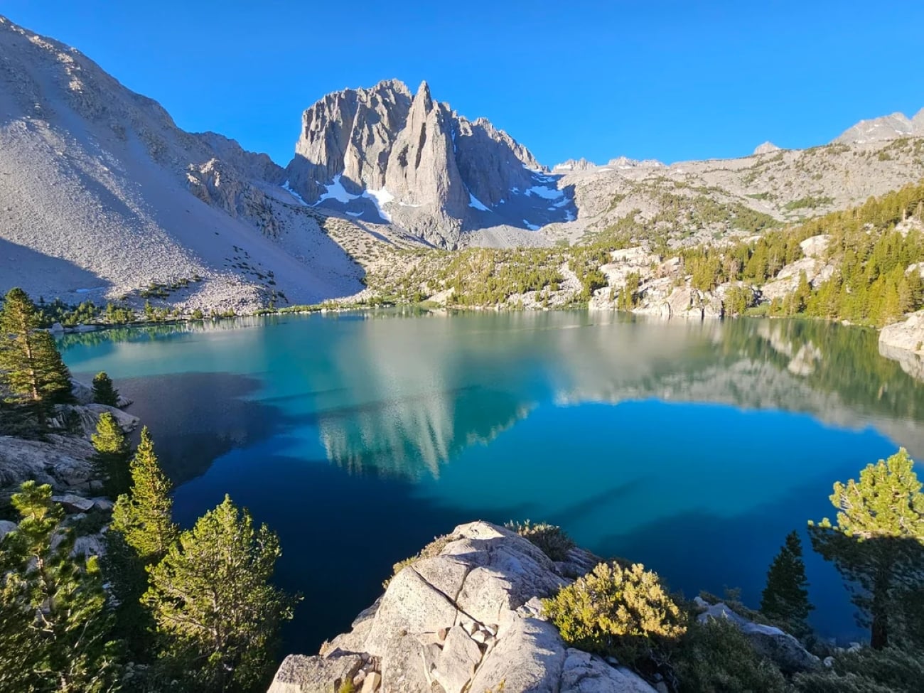 A clear blue lake reflects rugged mountains and a bright blue sky, surrounded by rocky slopes and scattered evergreen trees—a scene often posted by hikers in their travel photos.