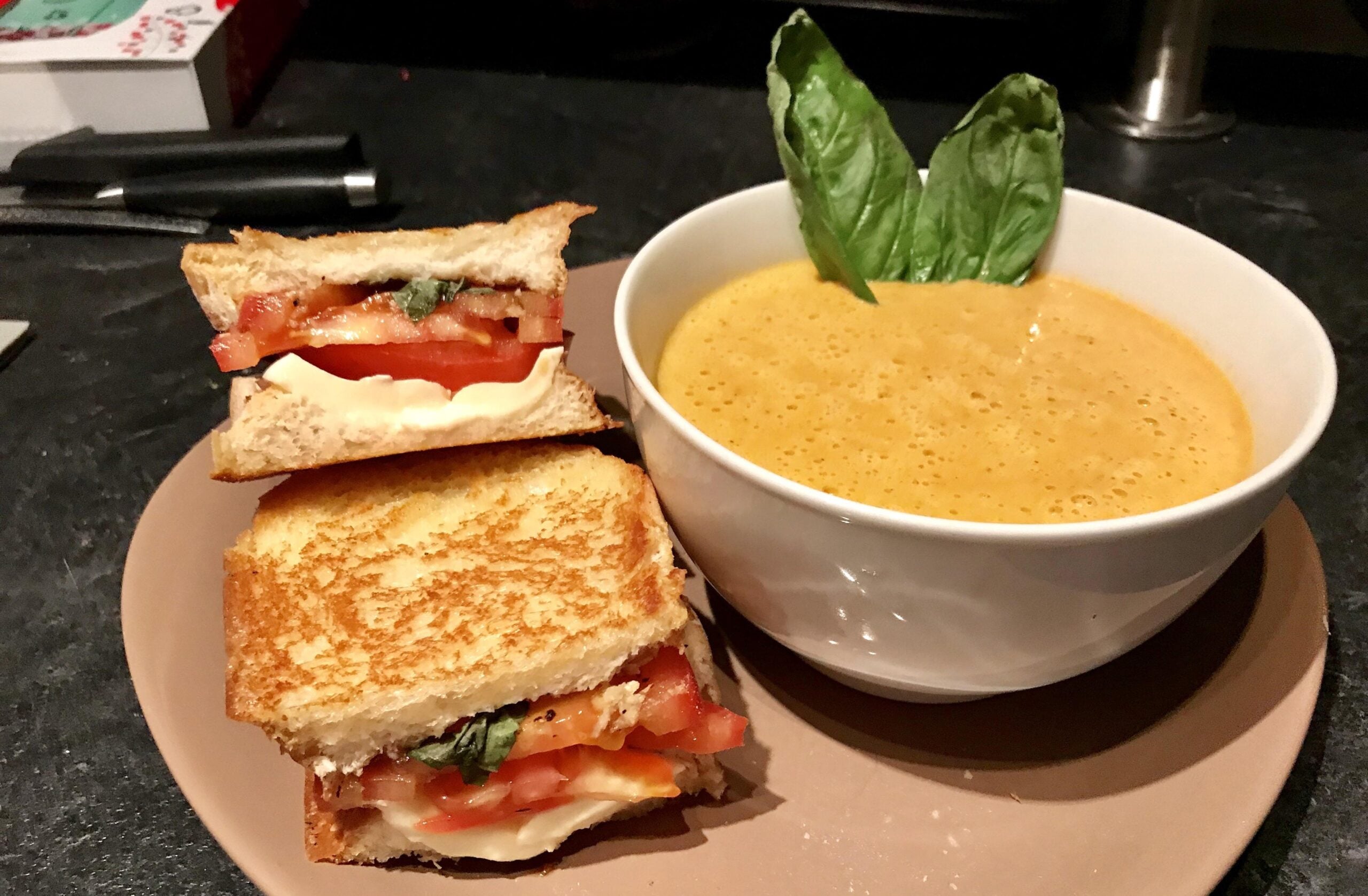A bowl of creamy tomato soup with two grilled cheese sandwiches and tomato, garnished with fresh basil leaves, on a beige plate.