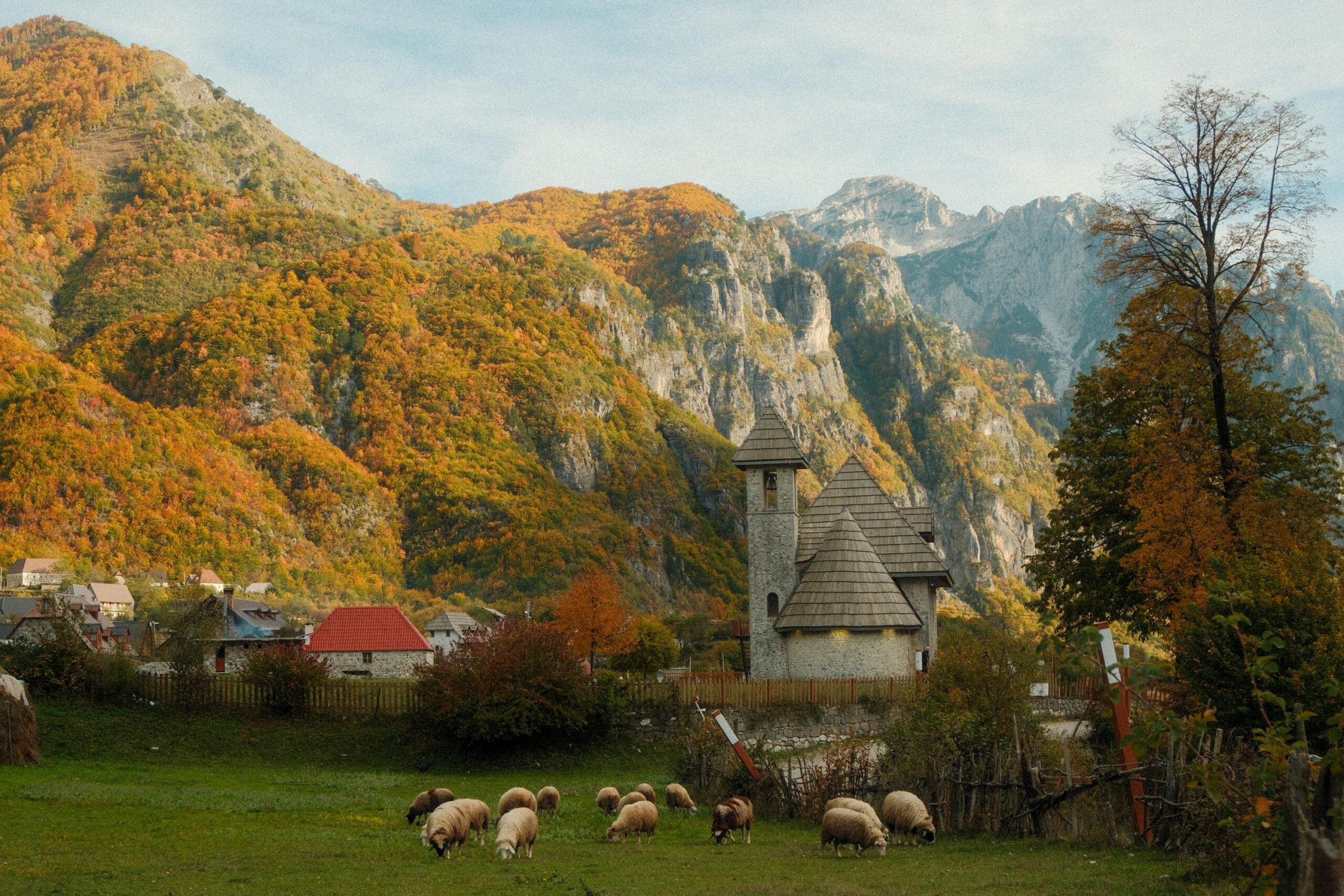 Stone church with pointed roofs stands in a grassy field with grazing sheep, surrounded by colorful autumn trees and dramatic, rocky mountains in the background.