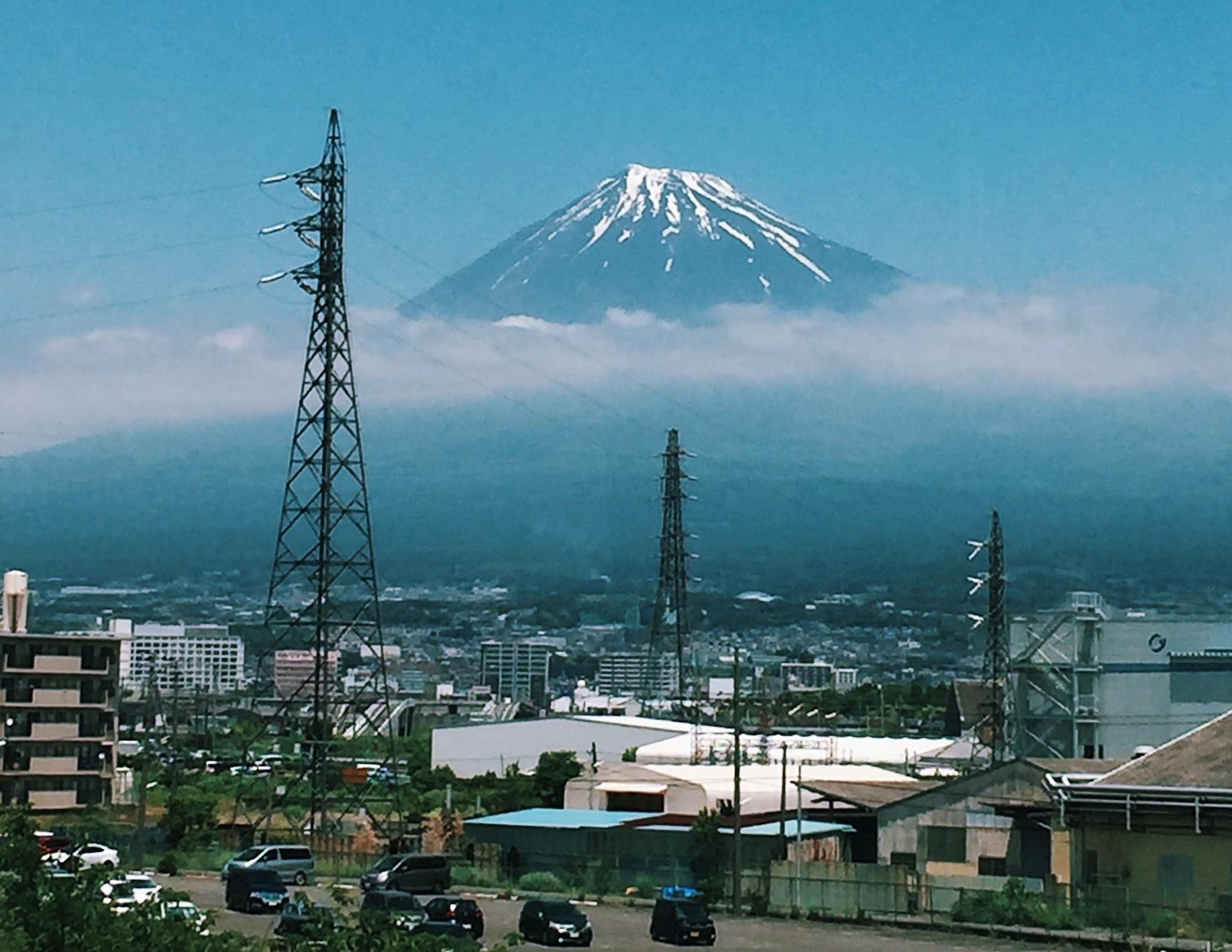 Snow-capped Mount Fuji rises above a cityscape with power lines and buildings in the foreground under a blue sky with scattered clouds.