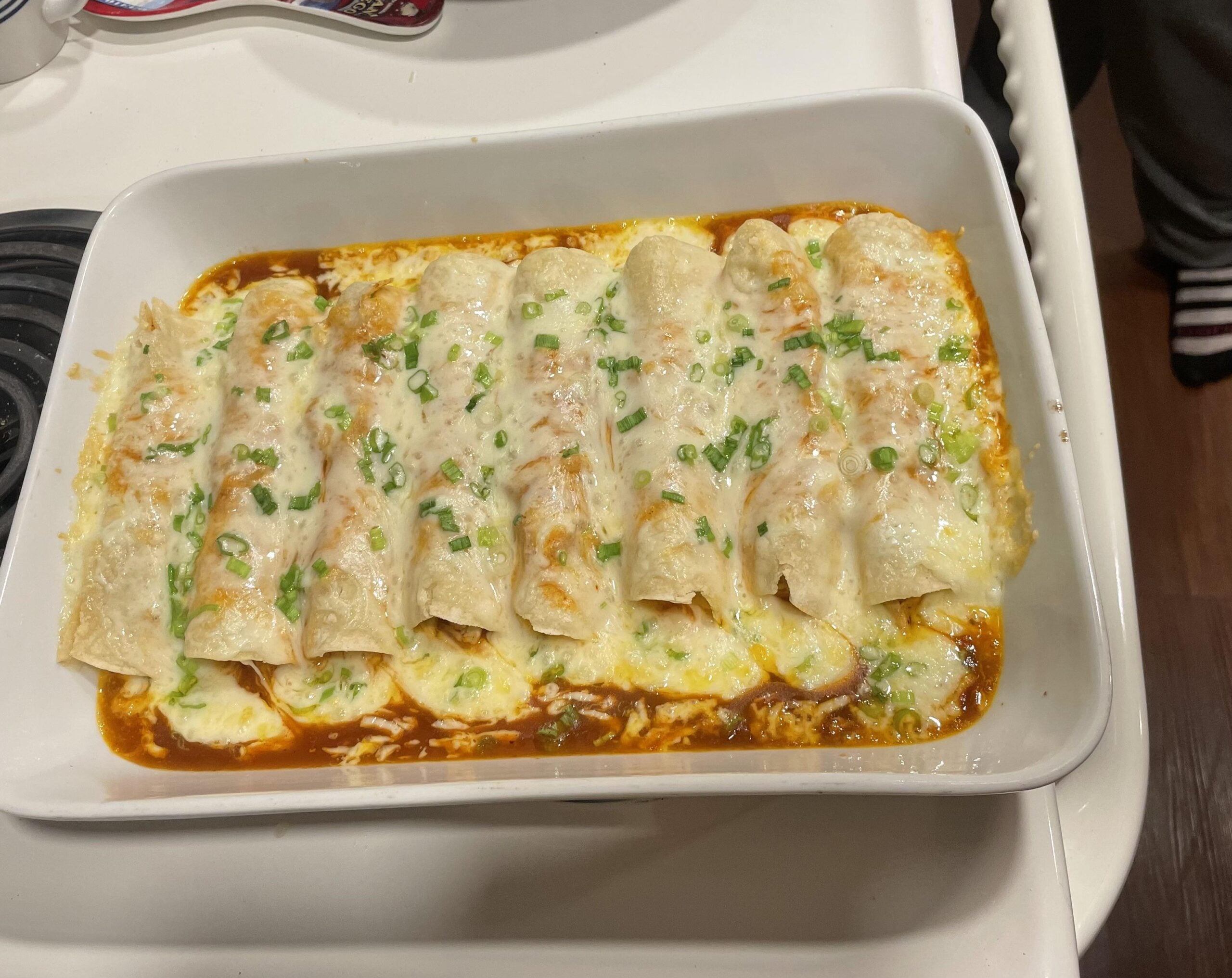 A white baking dish filled with eight cheese and green onion-topped enchiladas in red sauce, sitting on a white stove. Part of a person’s legs and striped socks are visible in the background.