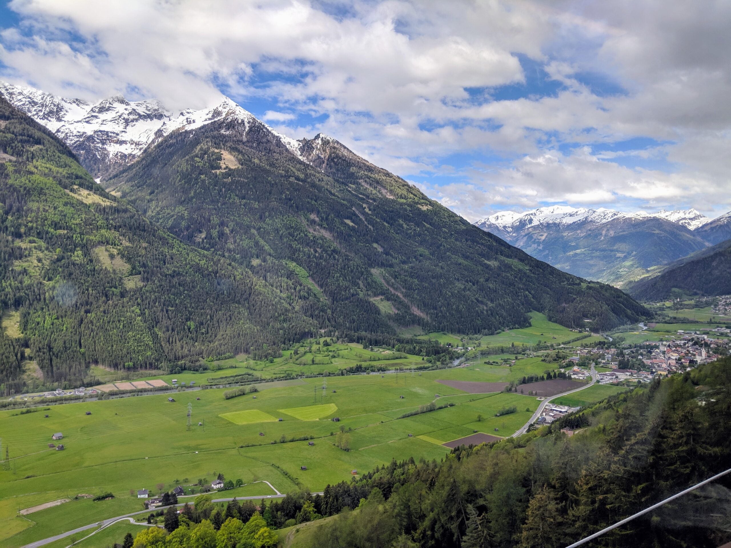 A scenic valley with green fields, a small town, and winding roads, surrounded by tall, snow-capped mountains under a partly cloudy sky.