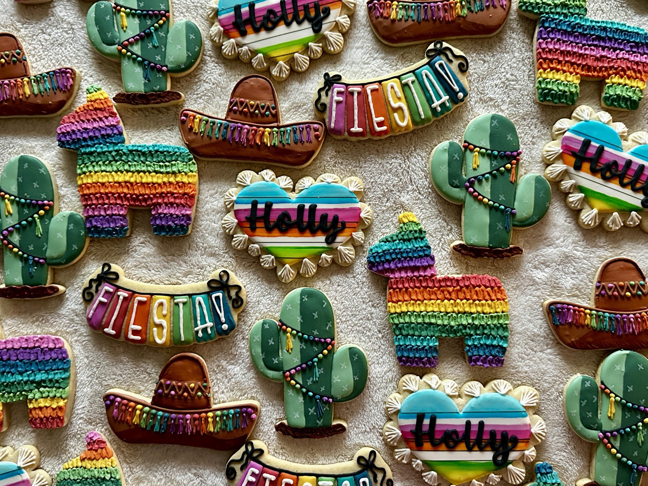 Colorful cookies shaped like cacti, piñatas, sombreros, hearts, and “Fiesta” banners, decorated with vibrant icing, are arranged on a textured light background. Some cookies have the name “Holly” written on them.