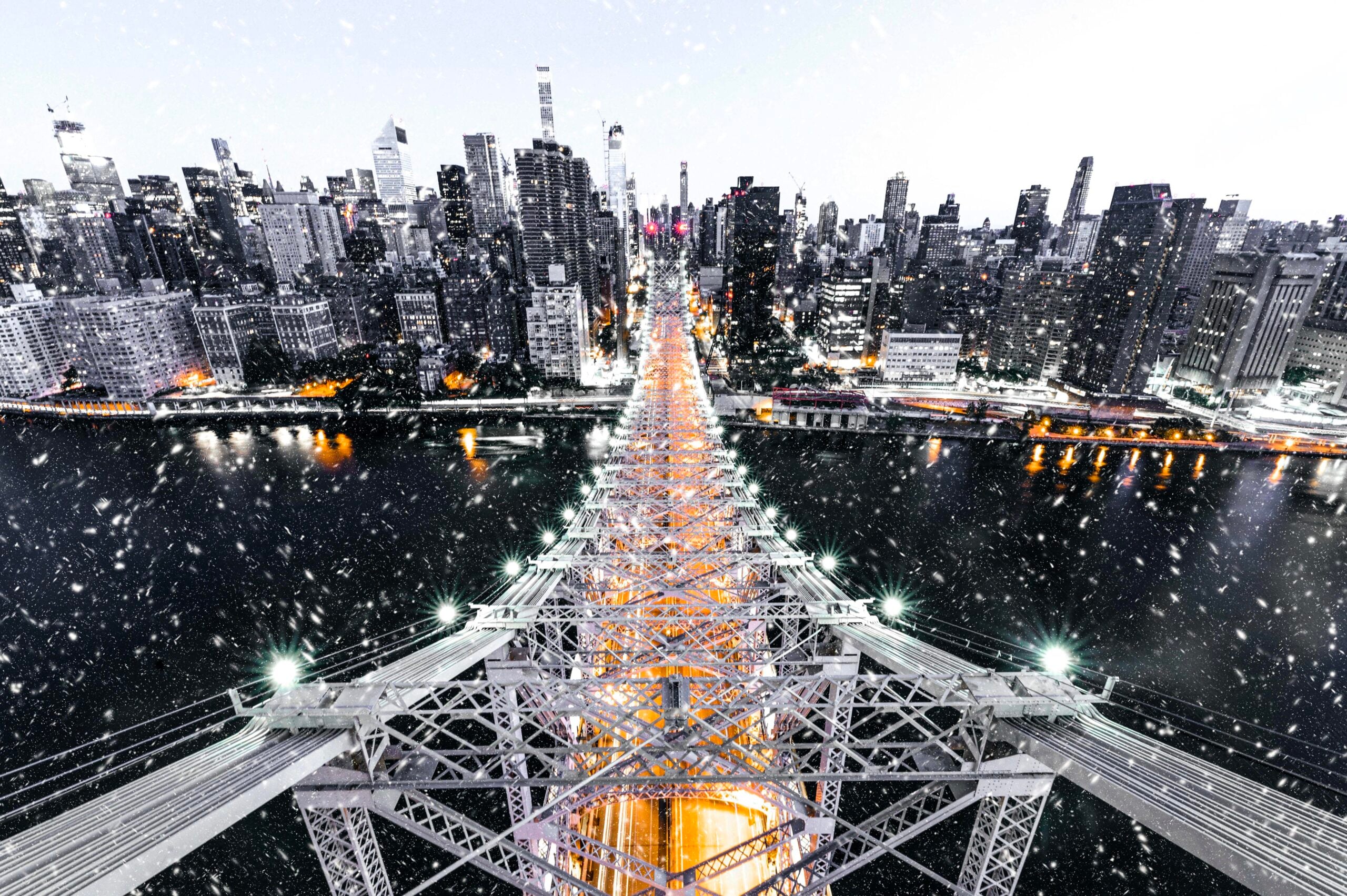 A snowy aerial view of a large suspension bridge leading into a city skyline with tall buildings, seen at dusk or dawn.