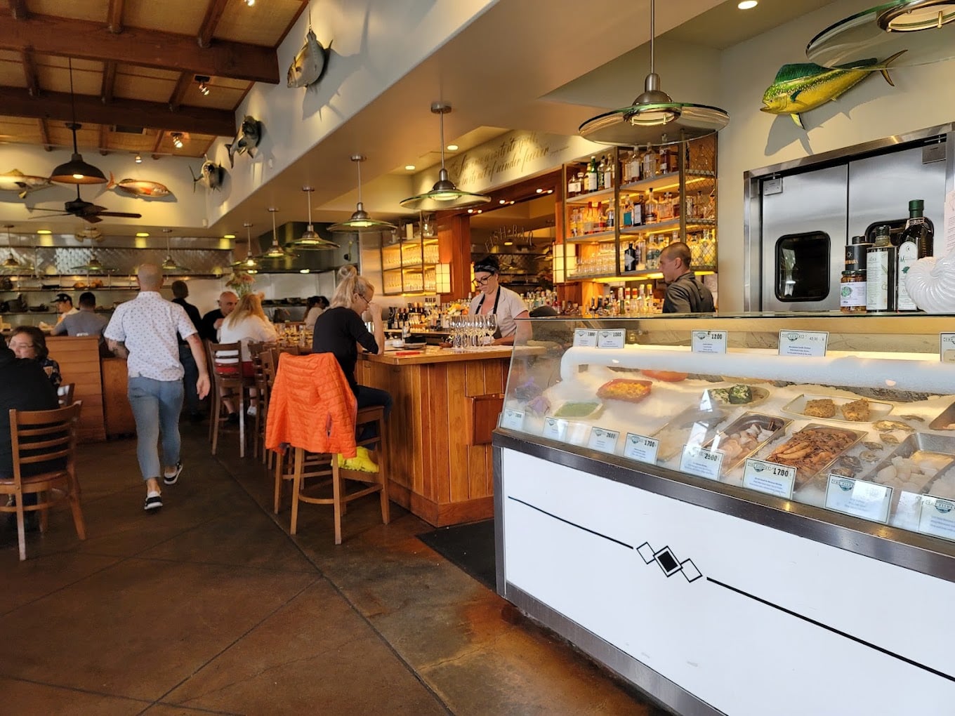 A seafood restaurant interior with customers at the bar, people dining, staff behind the counter, and a display case filled with fresh seafood. Mounted fish decorations are visible on the walls.