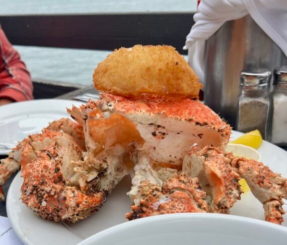 A cooked crab served on a white plate with a breaded fried item on top, accompanied by a lemon wedge. Salt and pepper shakers are visible in the background near a waterfront setting.