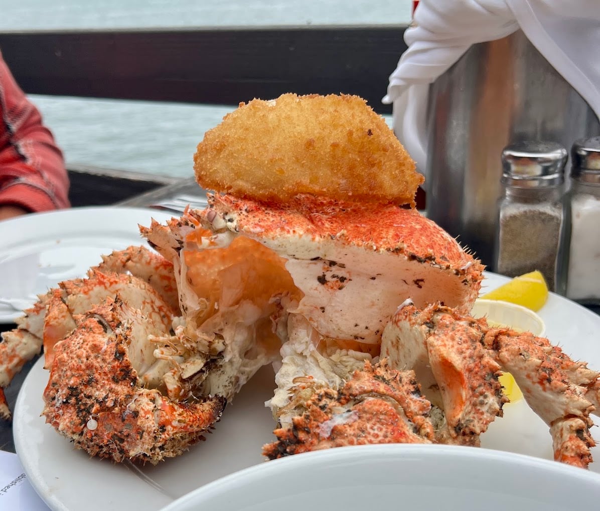 A cooked crab served on a white plate with a breaded fried item on top, accompanied by a lemon wedge. Salt and pepper shakers are visible in the background near a waterfront setting.