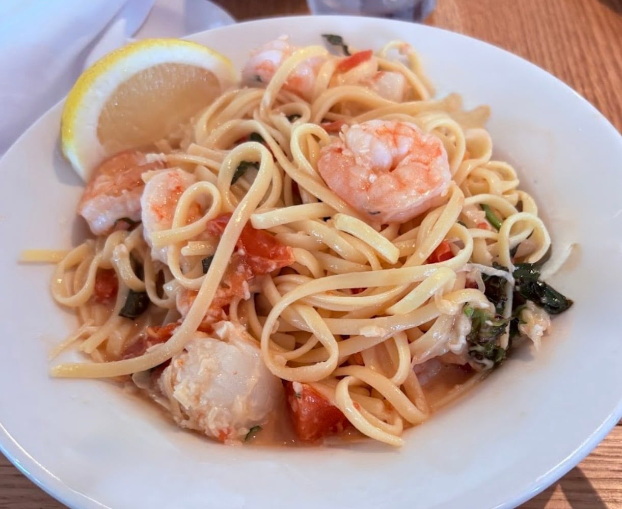 A white bowl filled with linguine pasta, shrimp, diced tomatoes, herbs, and a lemon wedge sits on a wooden table.