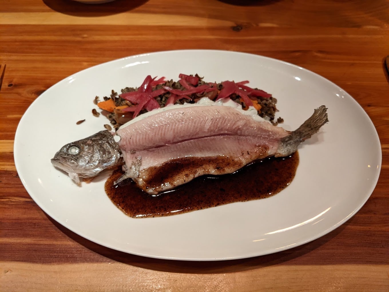 A whole cooked fish with white flesh is served on a white plate with dark sauce and a side of grains, carrots, and pickled vegetables on a wooden table.