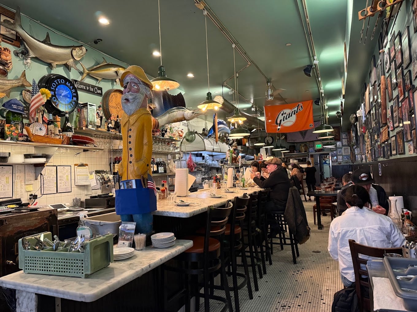 A busy seafood diner with a fisherman statue, mounted fish, and a Giants flag. People are eating at the long counter and tables, while staff work behind the counter and in the dining area.