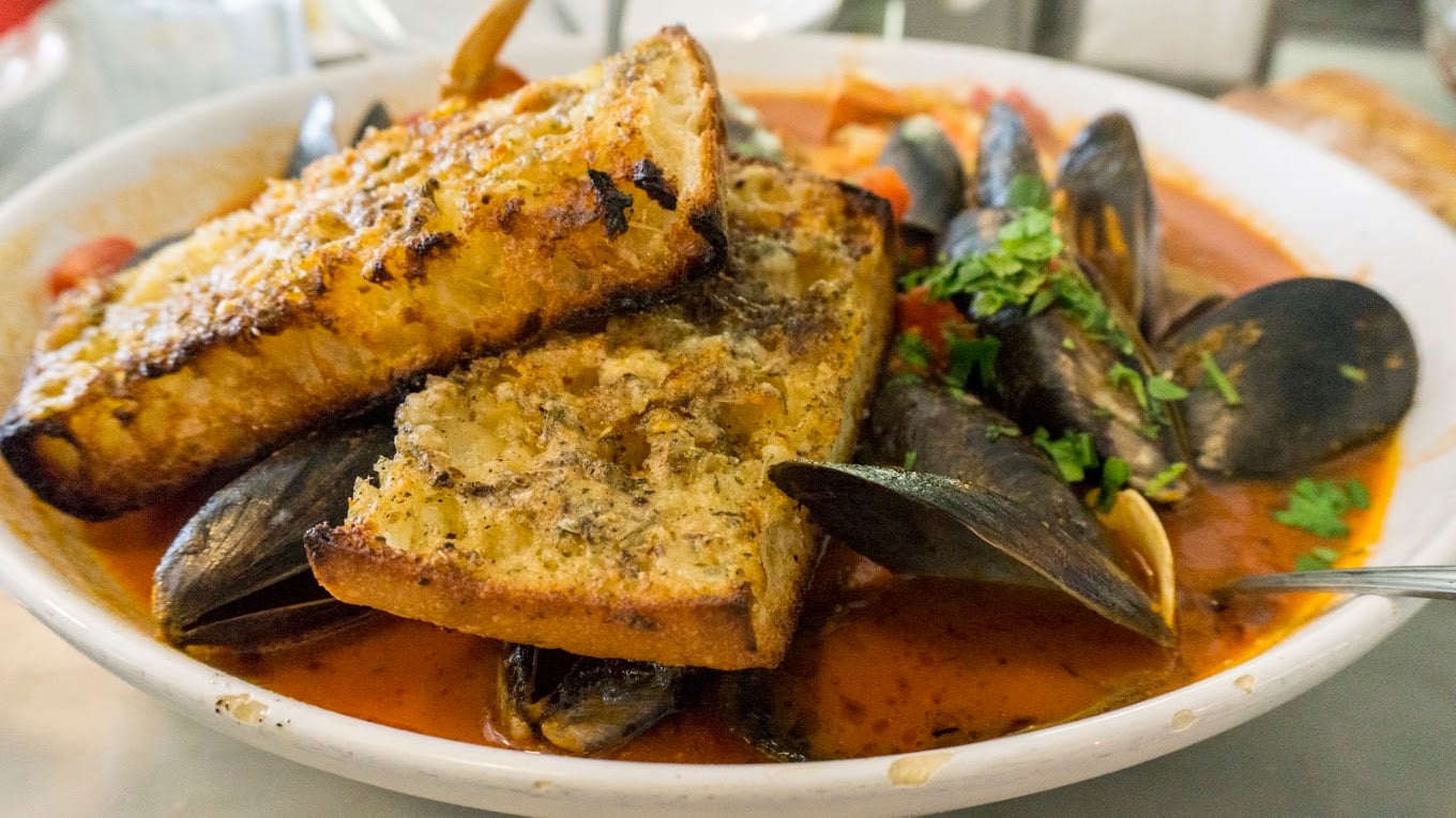 A bowl of seafood stew with mussels in tomato broth, topped with two slices of toasted bread and garnished with herbs.