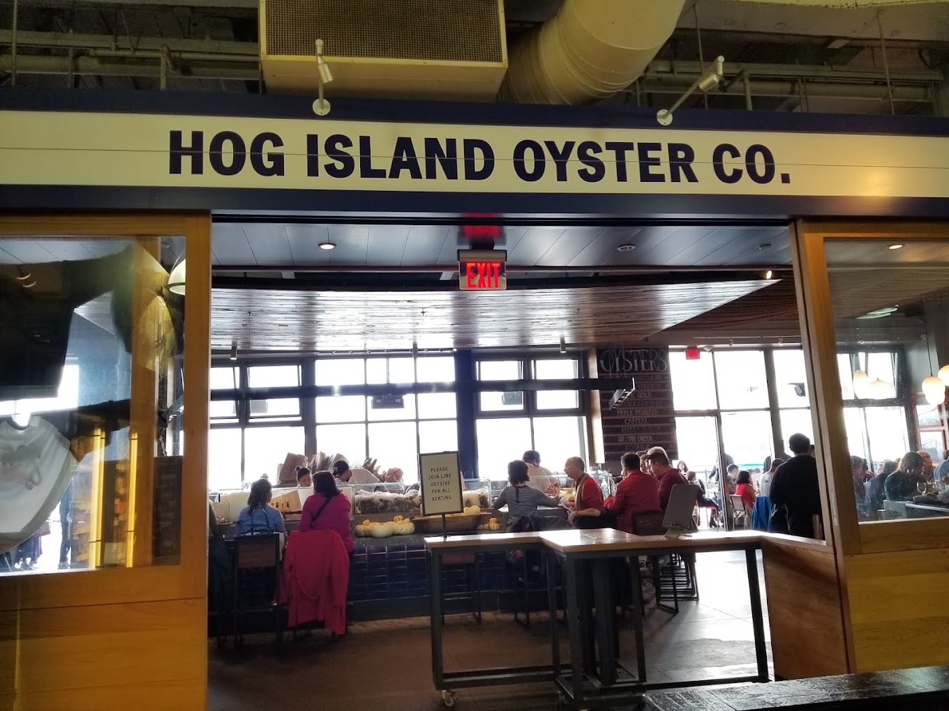 Large sign reads “Hog Island Oyster Co.” above a restaurant entrance, with people dining inside at tables and a bright window in the background.