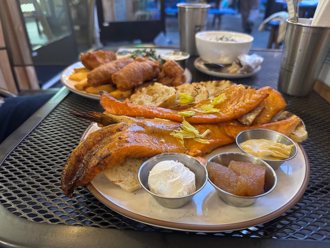A plate with smoked fish, bread slices, and sides of sour cream, mustard, and fruit compote sits on a metal table. In the background, there are fried foods and a bowl of soup.