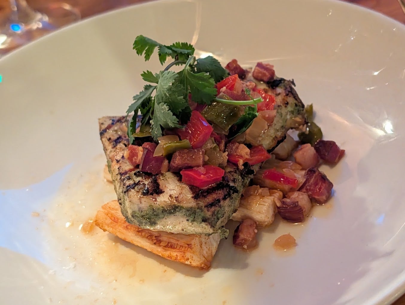 A grilled fish fillet topped with chopped vegetables and herbs sits on a white plate, garnished with cilantro and served over a root vegetable.