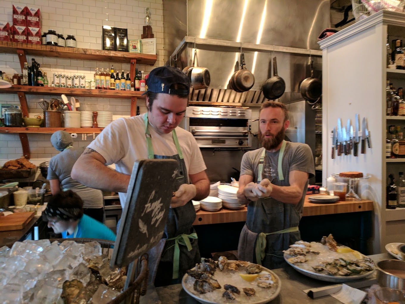 Two men in a restaurant kitchen shucking oysters behind a counter covered with ice and oysters; pots, pans, and shelves with bottles are seen in the background.