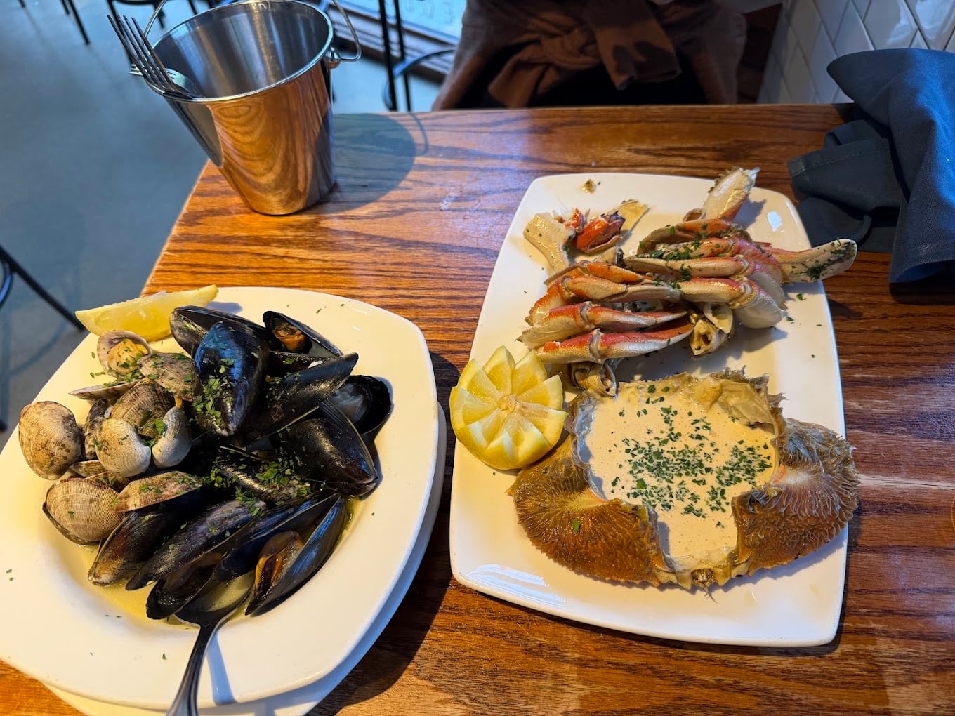 Two plates of seafood: one with steamed mussels and clams, the other with crab legs and a crab shell filled with sauce, both garnished with herbs. A lemon half and a metal bucket are also on the wooden table.